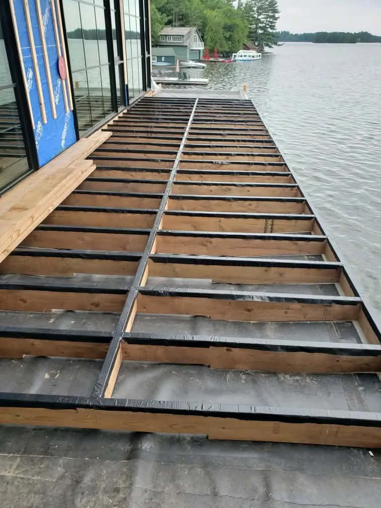 Wooden deck framing under construction along a lakeside with water and other houses in the background.