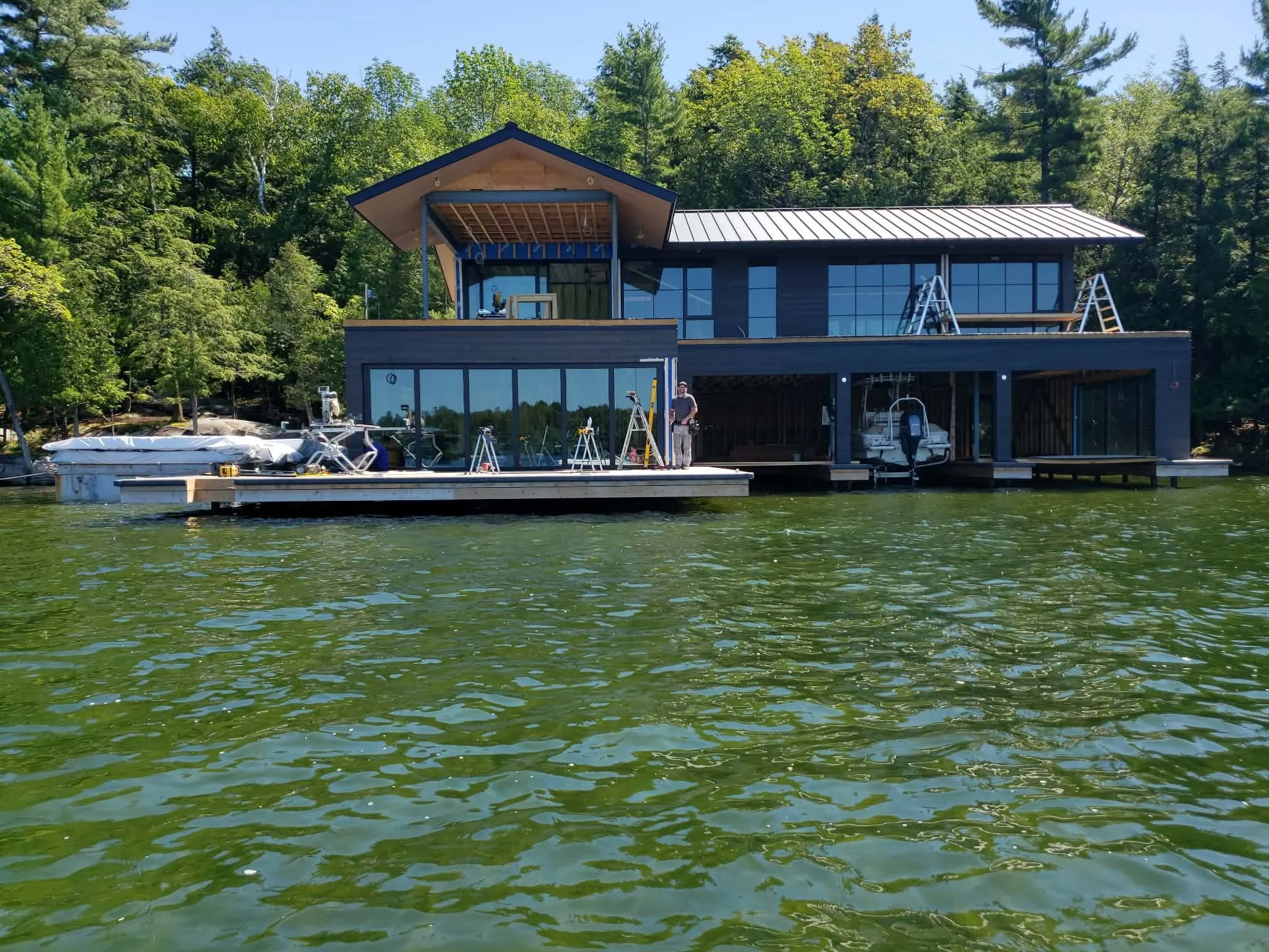 A modern two-story house under construction on a lake, with workers and ladders, surrounded by green trees.