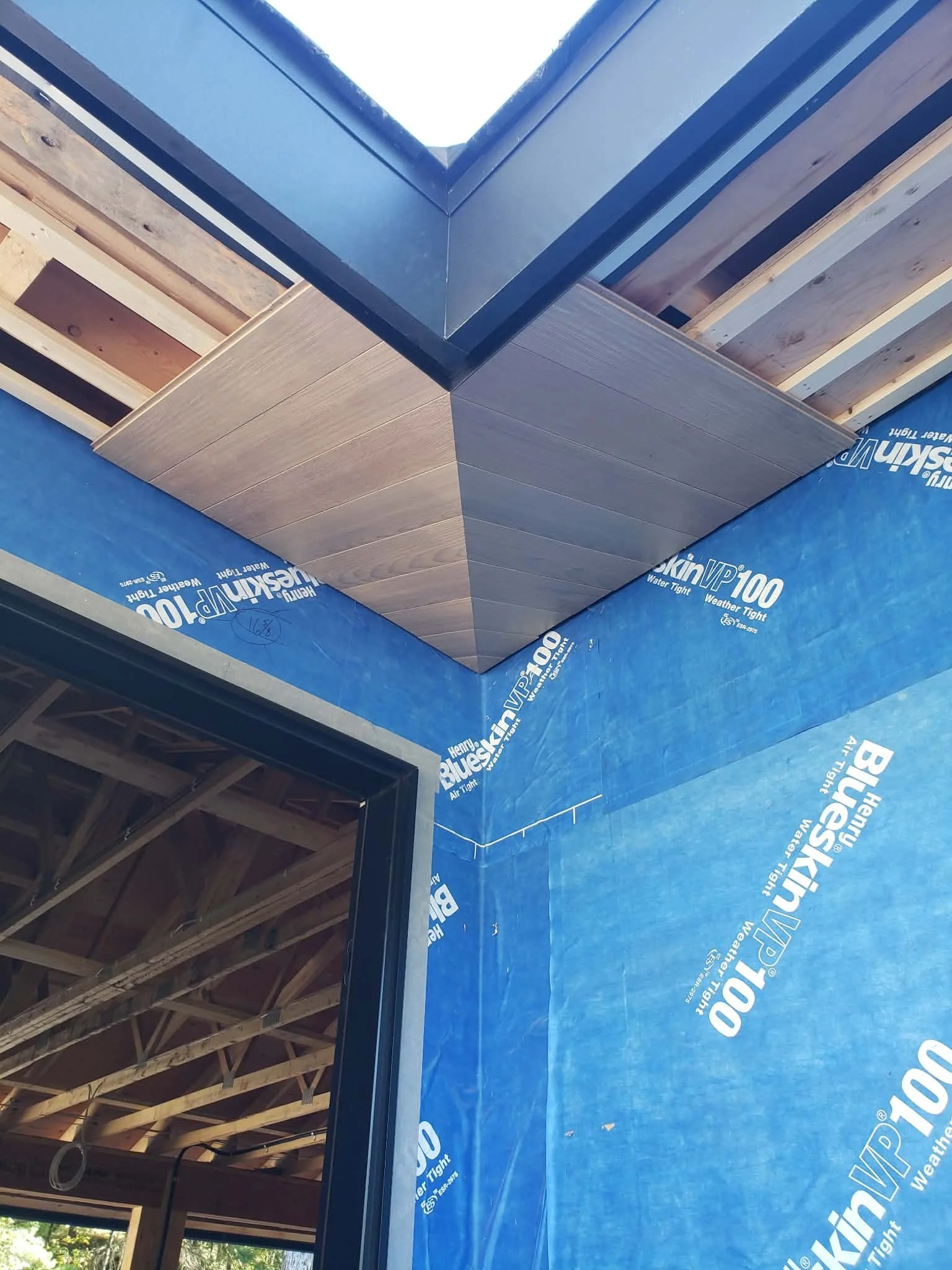 Corner of a building under construction showing blue Weather Barrier wrapping, wooden framing, and a black window frame.