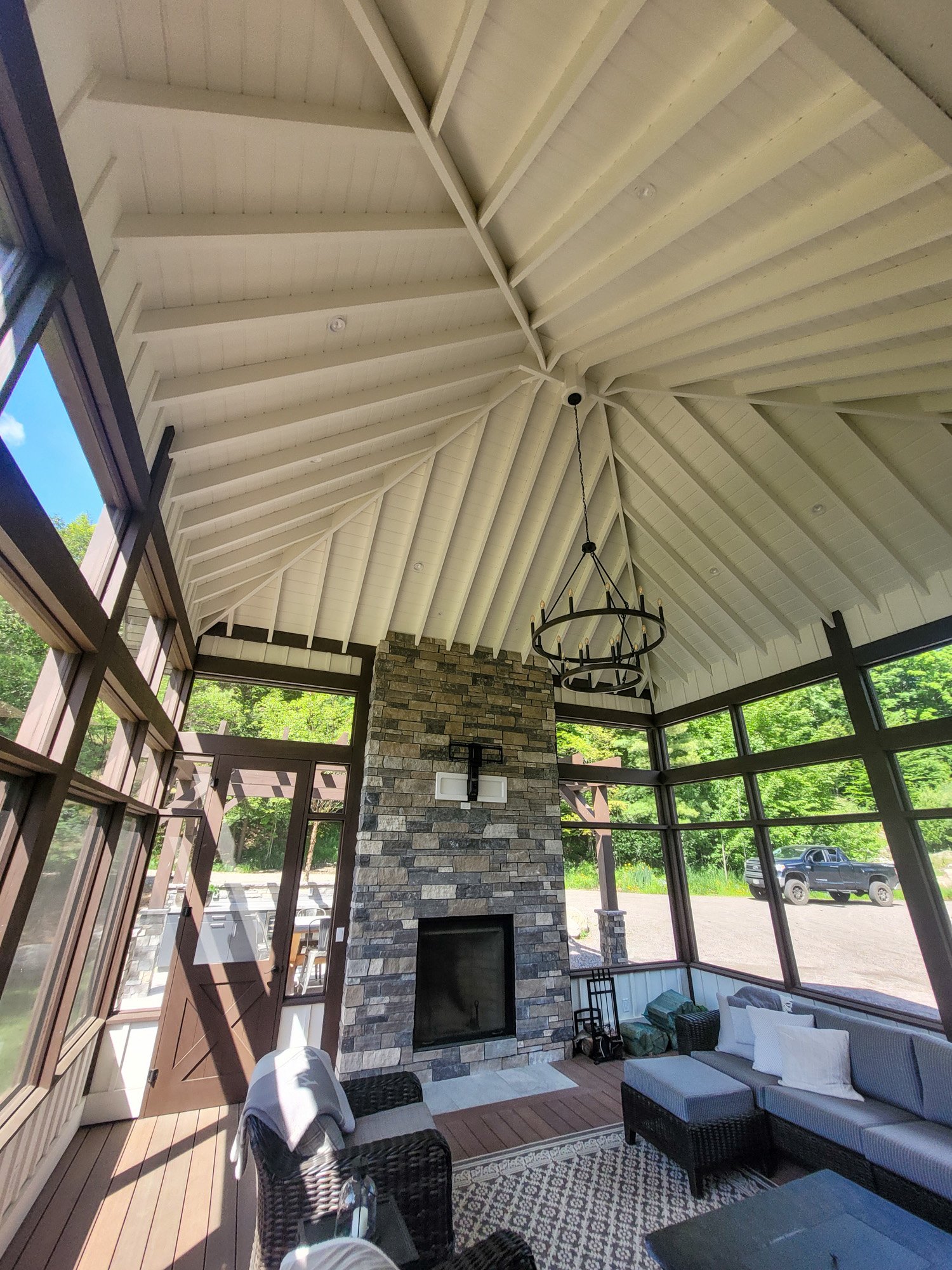 Sunroom with high, vaulted ceiling with exposed beams, a hanging chandelier, a stone fireplace, large windows with a view of a car and greenery outside, and outdoor furniture.