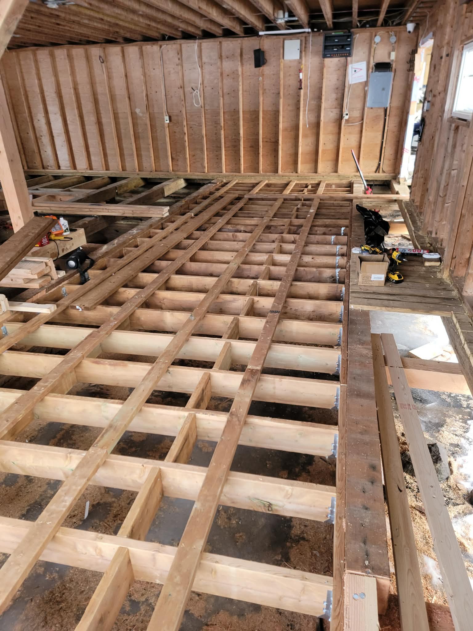 Interior of a house under construction with exposed wooden floor framing, tools, and construction materials visible.