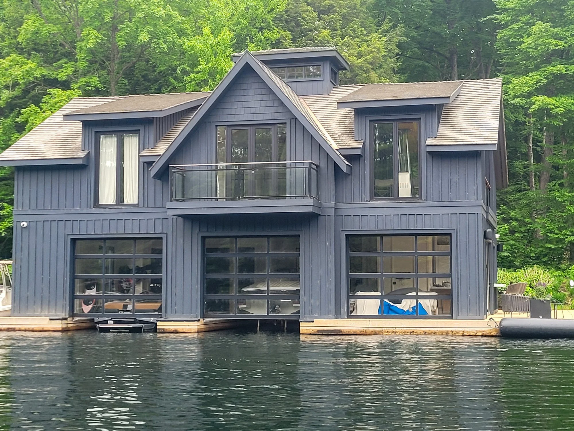 A blue wooden house with large glass garage doors is situated by a body of water, with a forested hillside in the background. The house features a balcony and multiple windows.