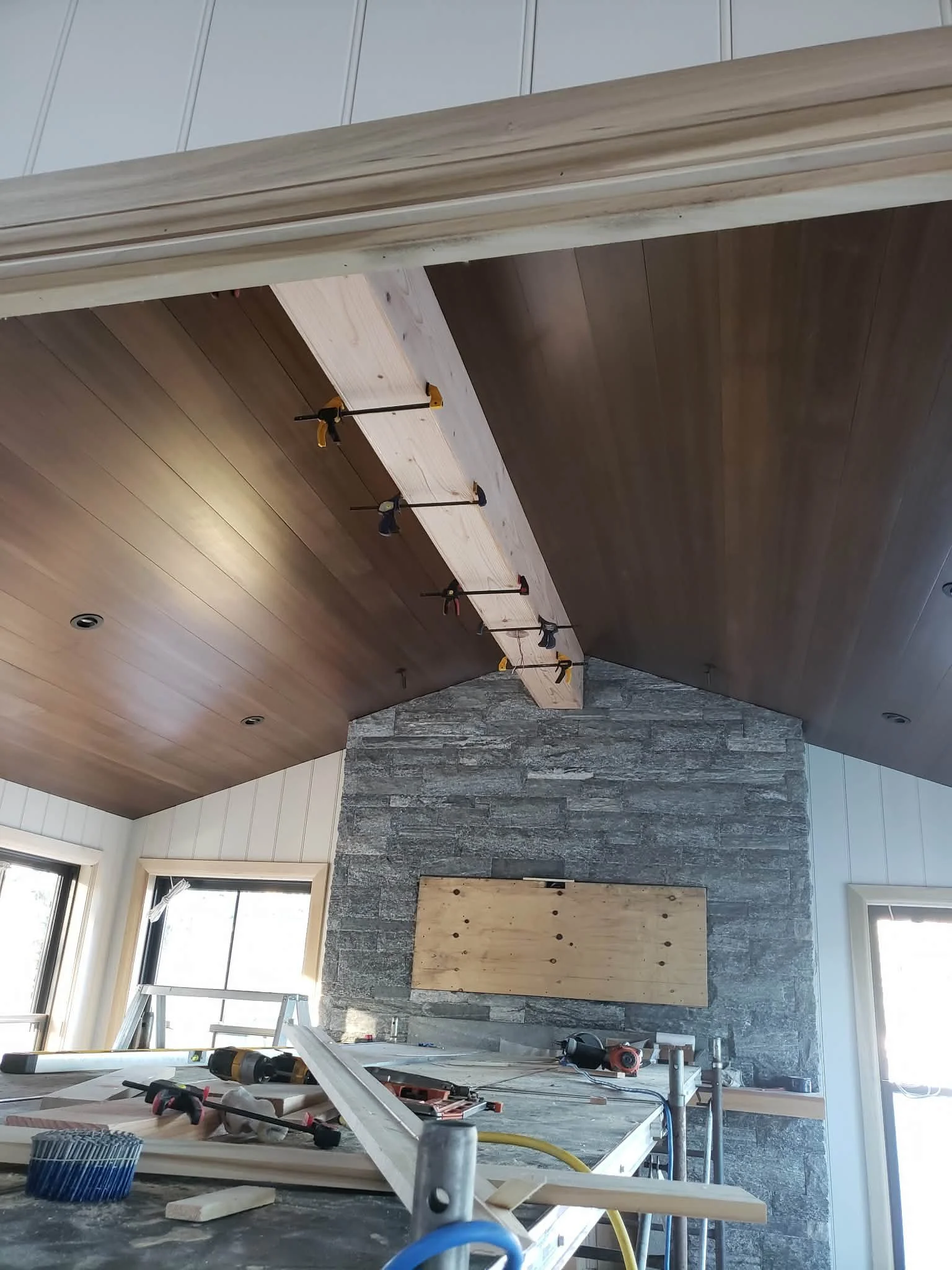 Interior of a room under construction, with a high vaulted ceiling featuring wood paneling, a stone fireplace, and window frames. Construction tools and materials are scattered on a work table in the foreground.