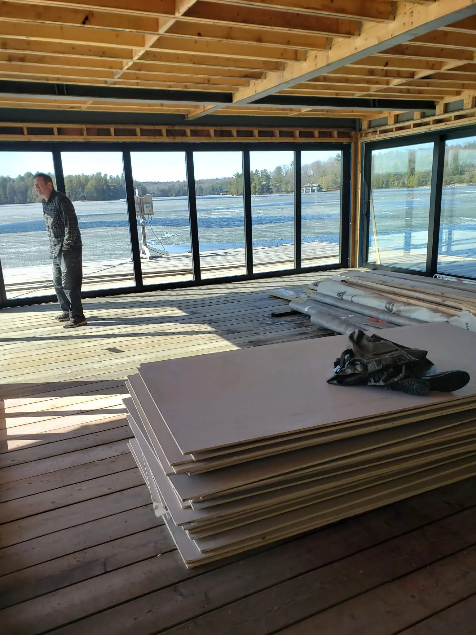 Interior view of a house under construction, showing wooden flooring, framed ceiling, large windows overlooking a snowy landscape with trees, stacks of drywall and construction tools inside.
