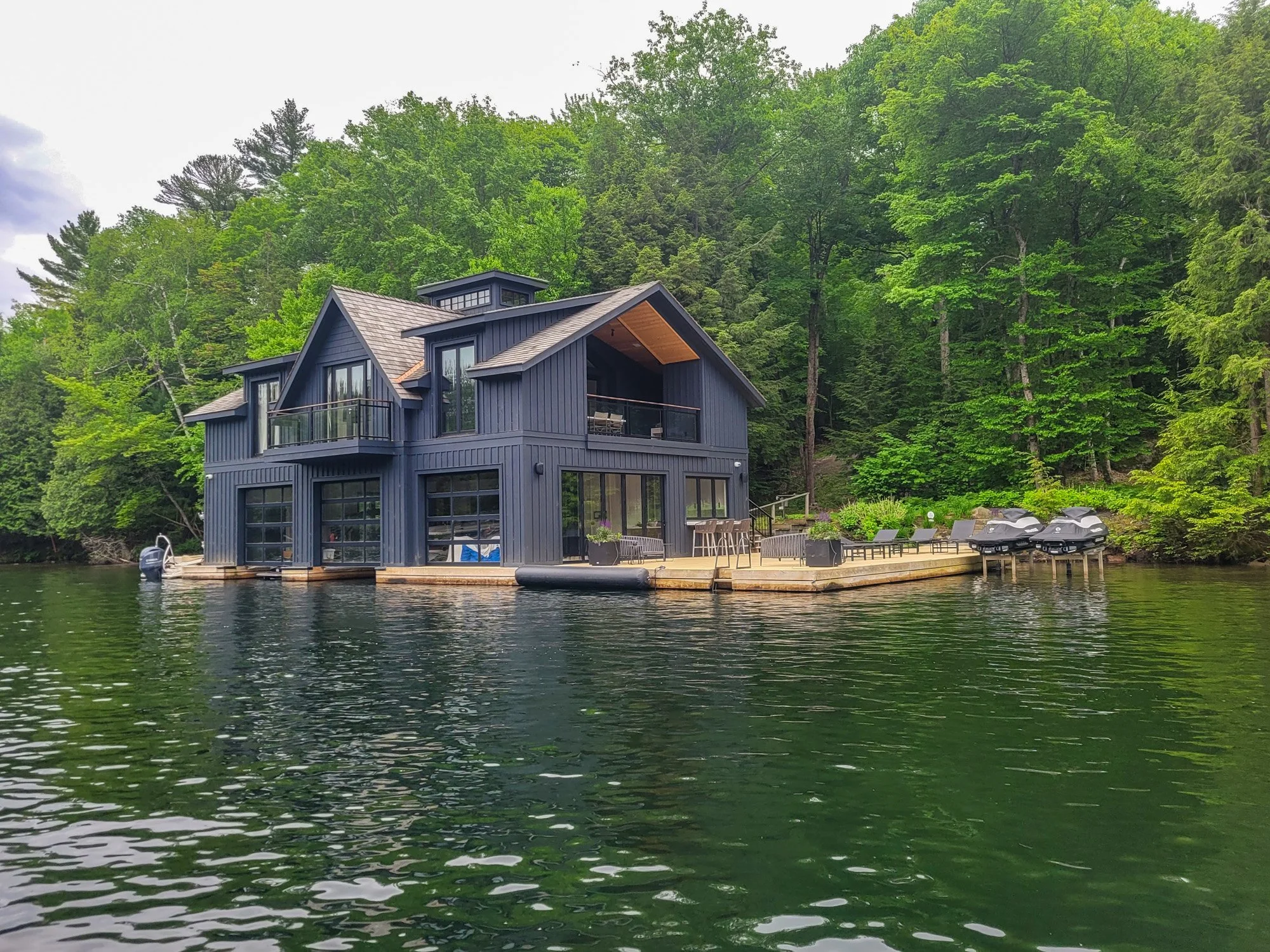 Modern blue house on a lake with a deck, outdoor furniture, and four jet skis, surrounded by green trees.