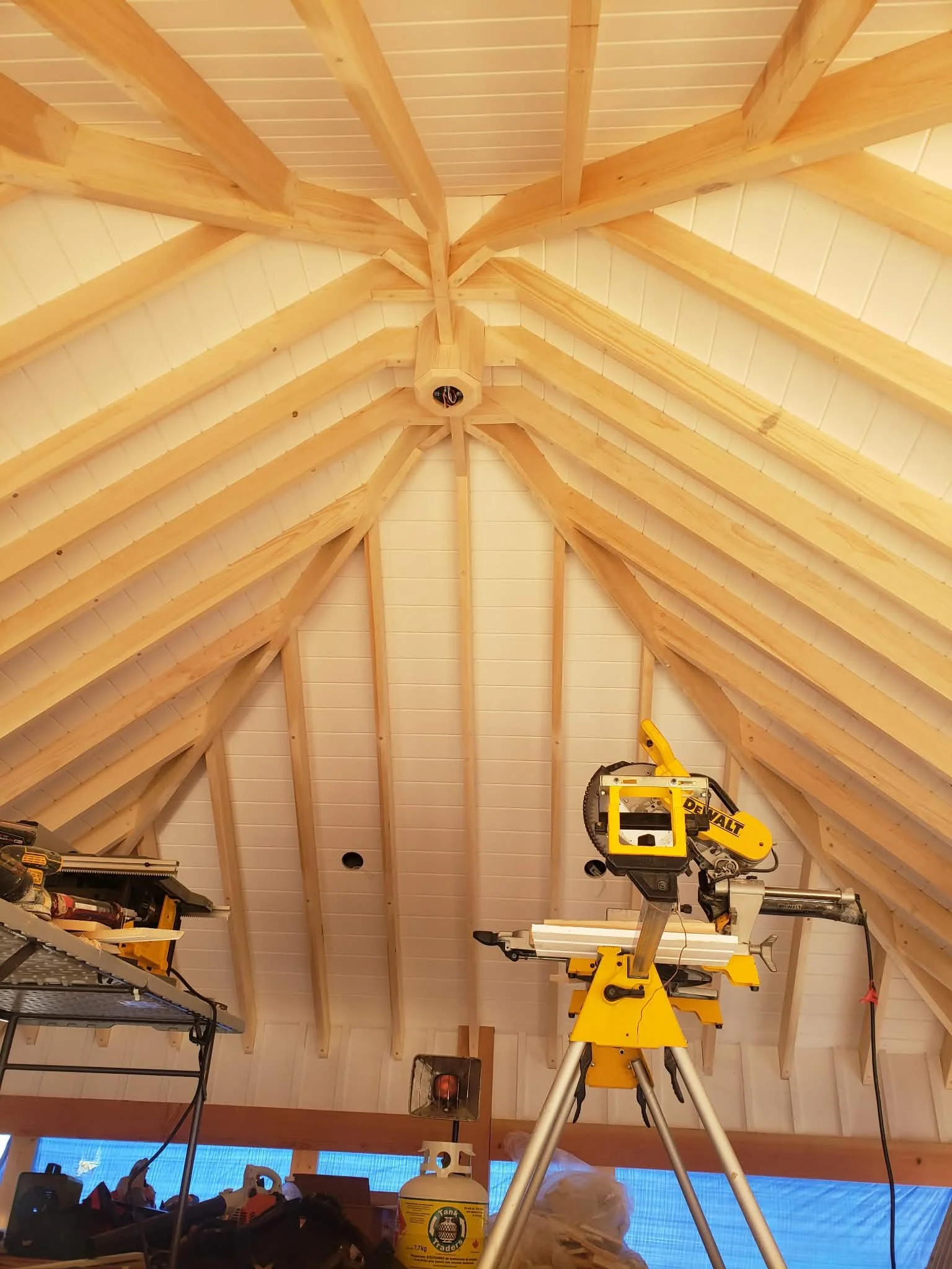 Interior view of a building under construction, showing wooden roof trusses and a yellow DeWalt miter saw on a stand.