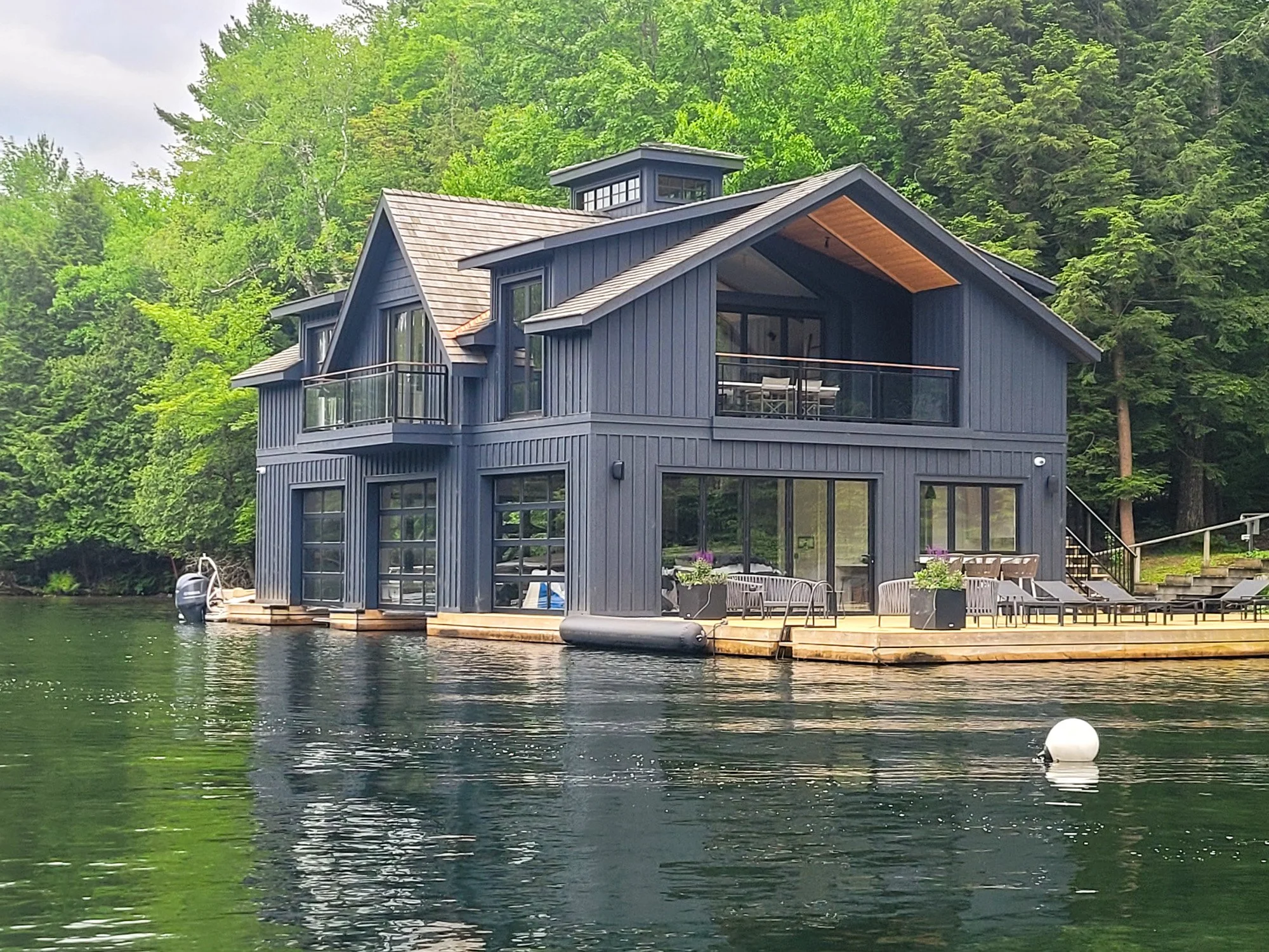 A modern two-story house painted dark blue is situated on the edge of a lake, with a wooden deck extending over the water. The house features large glass windows and balconies, surrounded by green trees.