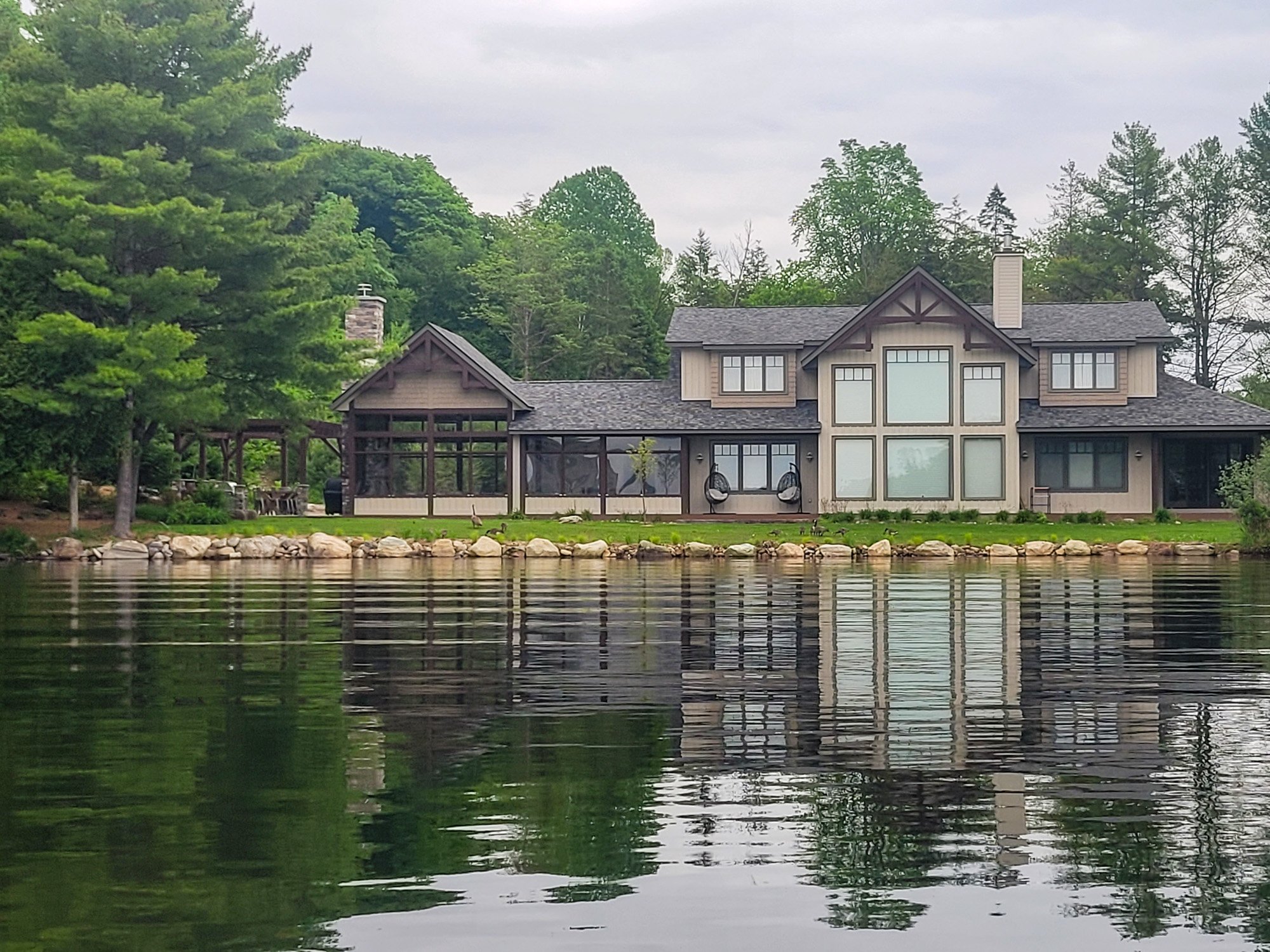 A modern house with large windows is situated by a body of water, surrounded by trees, with a rocky shoreline and a cloudy sky.