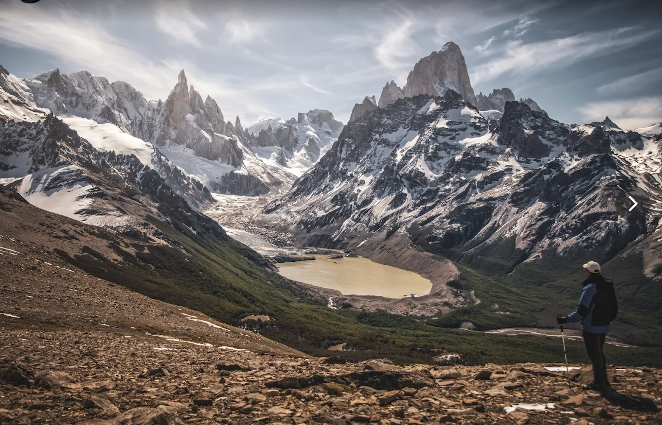 Una persona con equipo de senderismo observando un paisaje montañoso con picos nevados, un lago en el valle y un cielo parcialmente nublado.