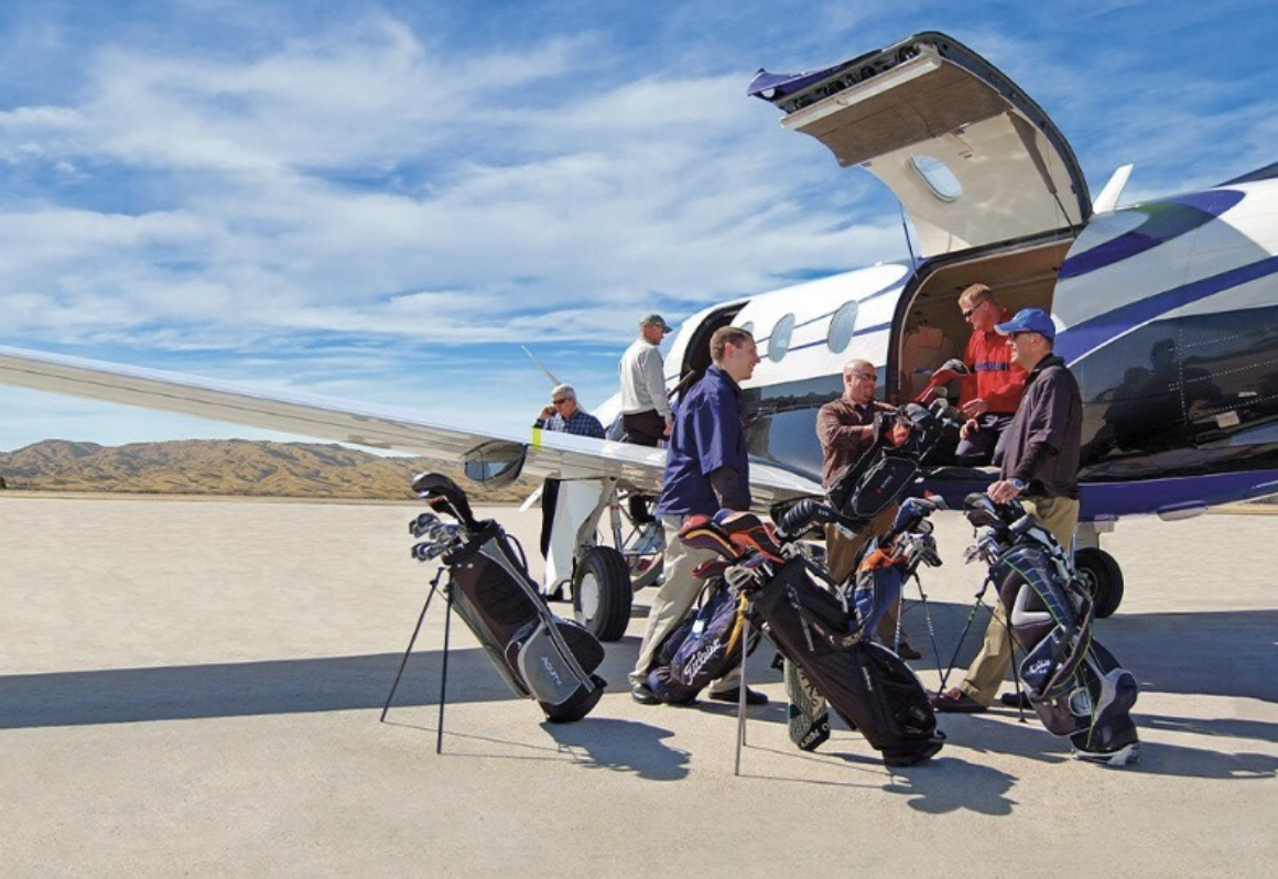 Grupo de golfistas con bolsas de golf junto a un avión en una pista y un paisaje montañoso en el fondo.