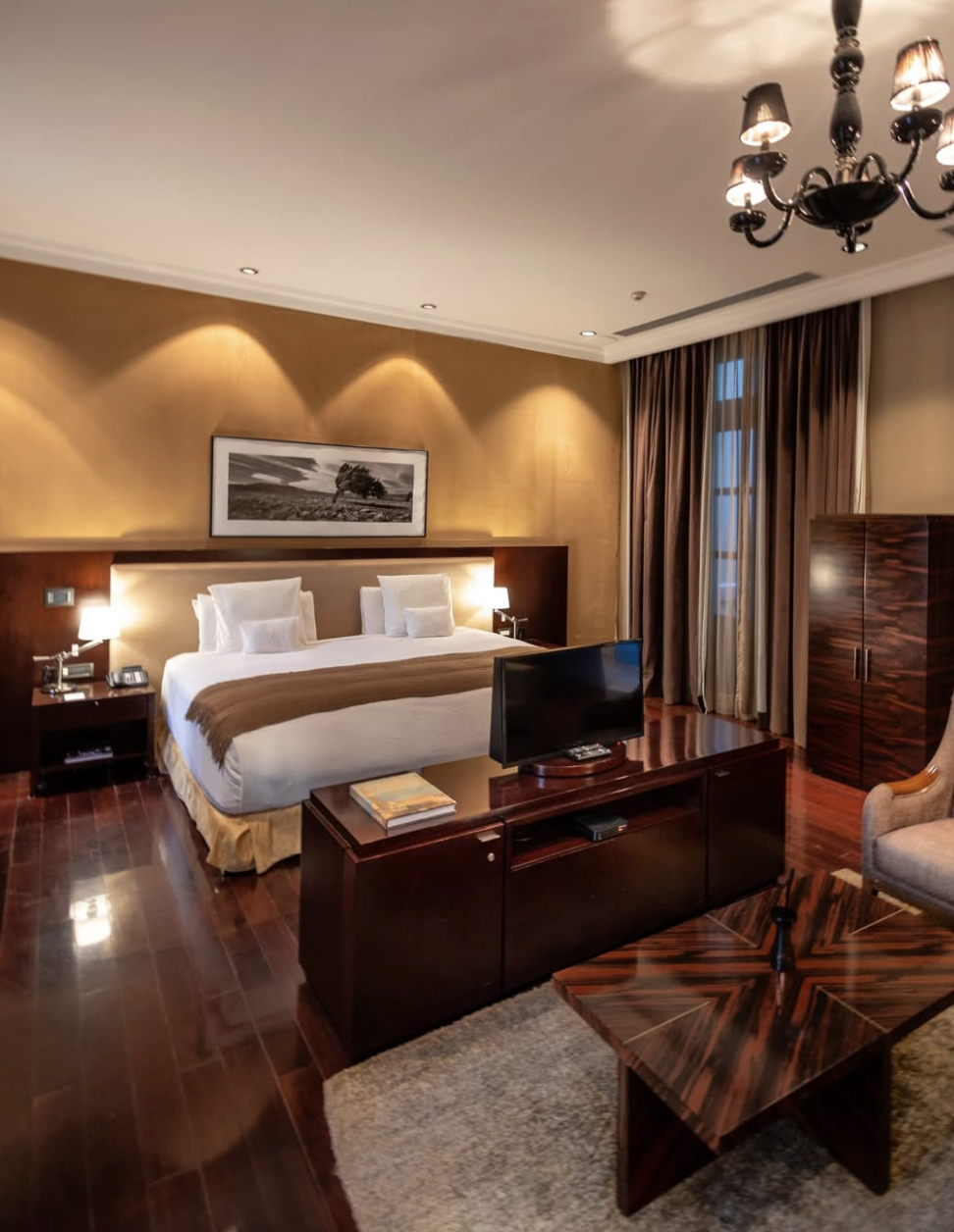 Habitación de hotel elegante con cama grande, cortinas largas, mobiliario de madera oscura y televisión en un mueble, decorada con un cuadro y iluminación cálida.