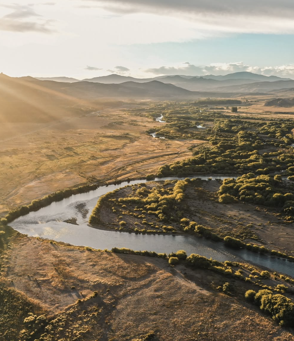 Vista aérea de un río serpenteante en un paisaje de llanuras y colinas con vegetación dispersa, bajo un cielo parcialmente nublado con luz solar en el horizonte.
