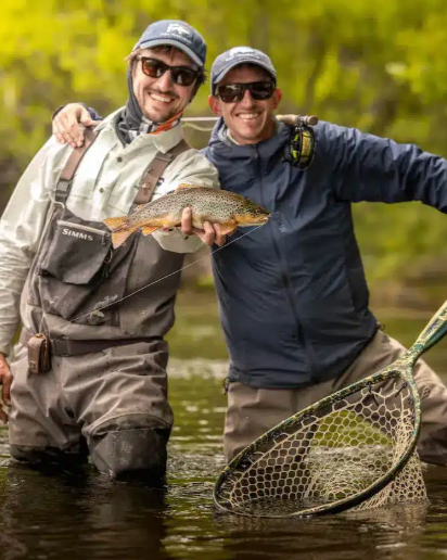 Dos hombres sonriendo en un río, sosteniendo una trucha pescada, con una red de pesca y un entorno natural con árboles.