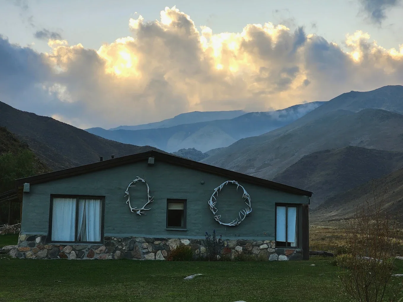 Casa de campo en un valle con montañas de fondo y cielos nublados con sol.