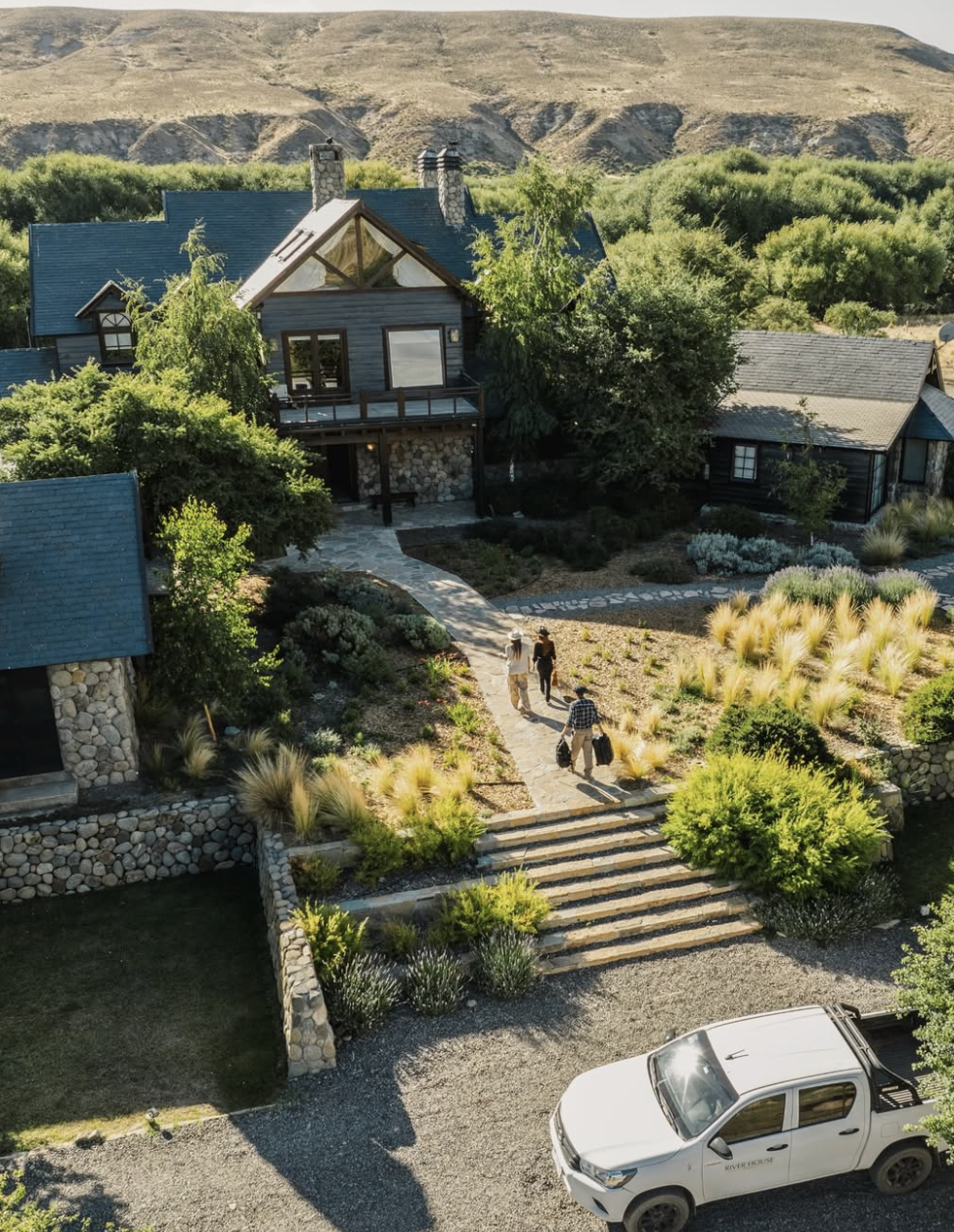 Casa de estilo rústico con techos azules y chimeneas, rodeada de vegetación y caminos de piedra, con personas caminando y un coche parked.