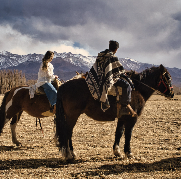 Dos personas montando caballos en un campo con montañas y cielo nublado en el fondo.