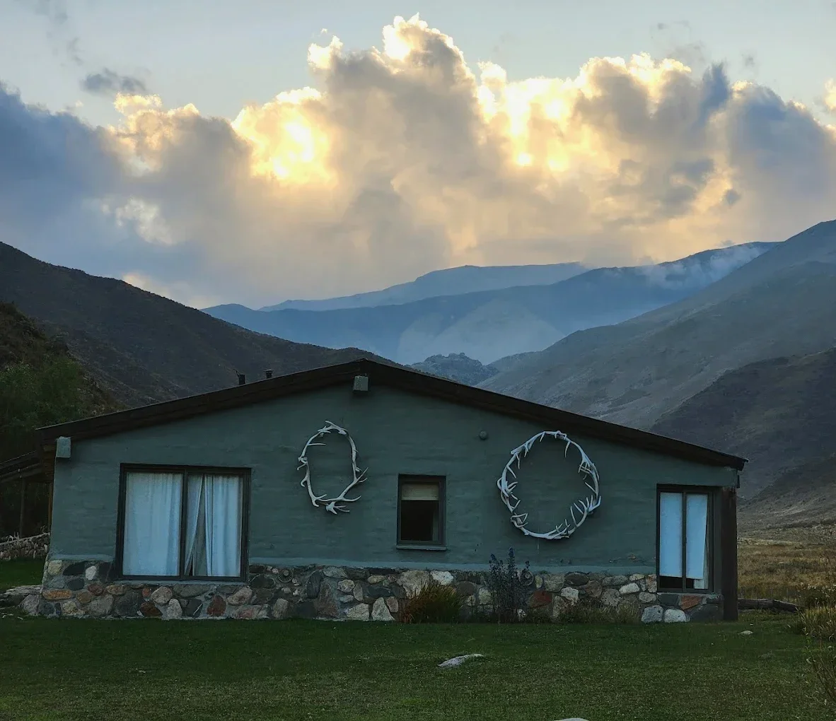 Casa de piedra con ventanas y decoraciones en forma de círculos de ramas, ubicada en un paisaje montañoso con nubes y cielo al atardecer.