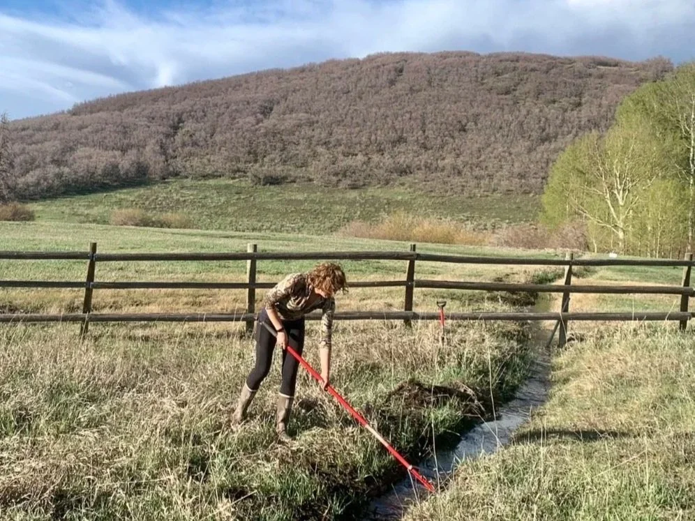 A woman with long curly hair, wearing rain boots and dark pants, cleaning a small creek with a red rake in a grassy field, with a wooden fence and rolling hills in the background.