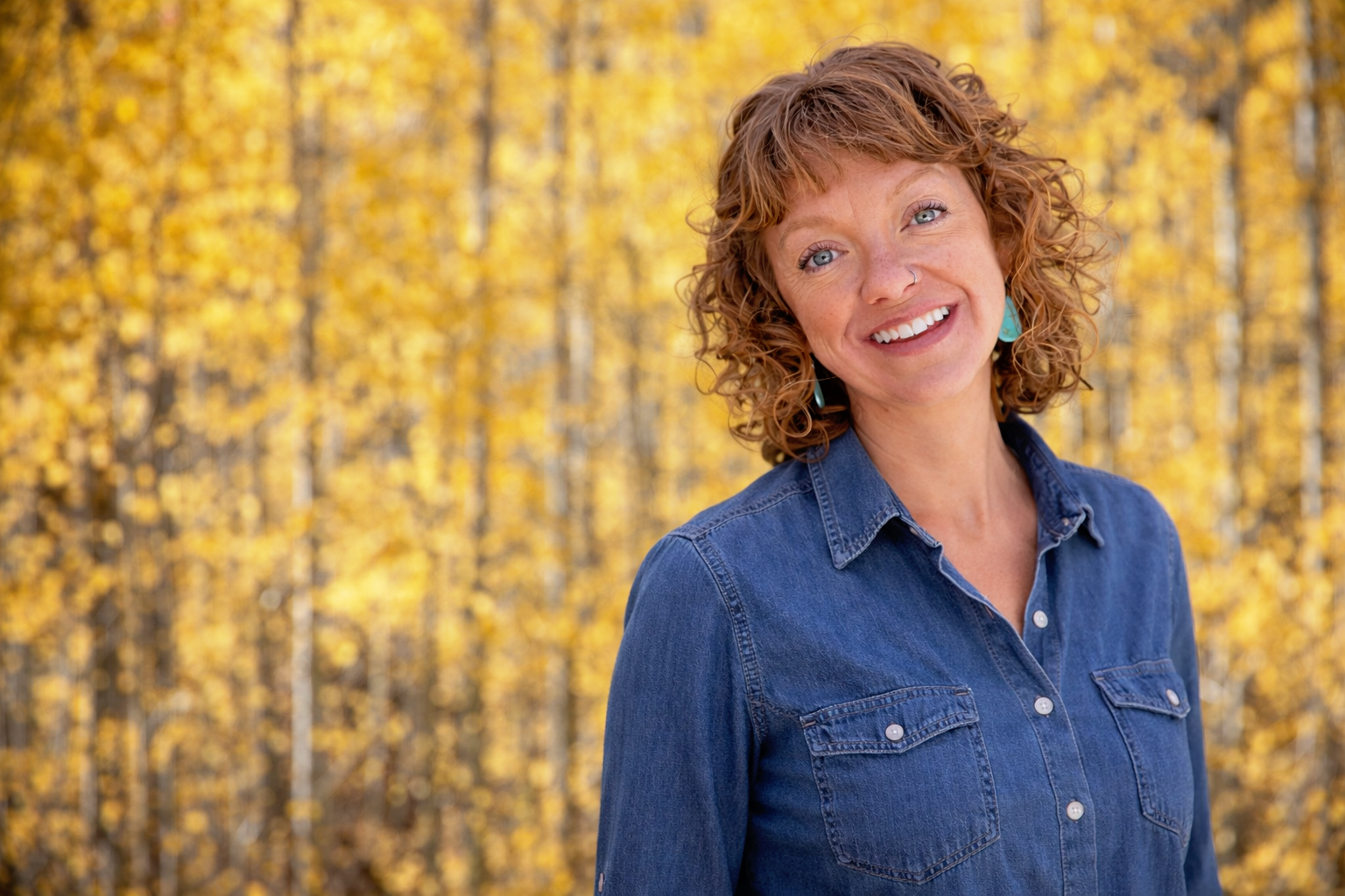 A woman with curly red hair and blue eyes smiling outdoors in front of yellow autumn leaves, wearing a denim shirt and turquoise earrings.