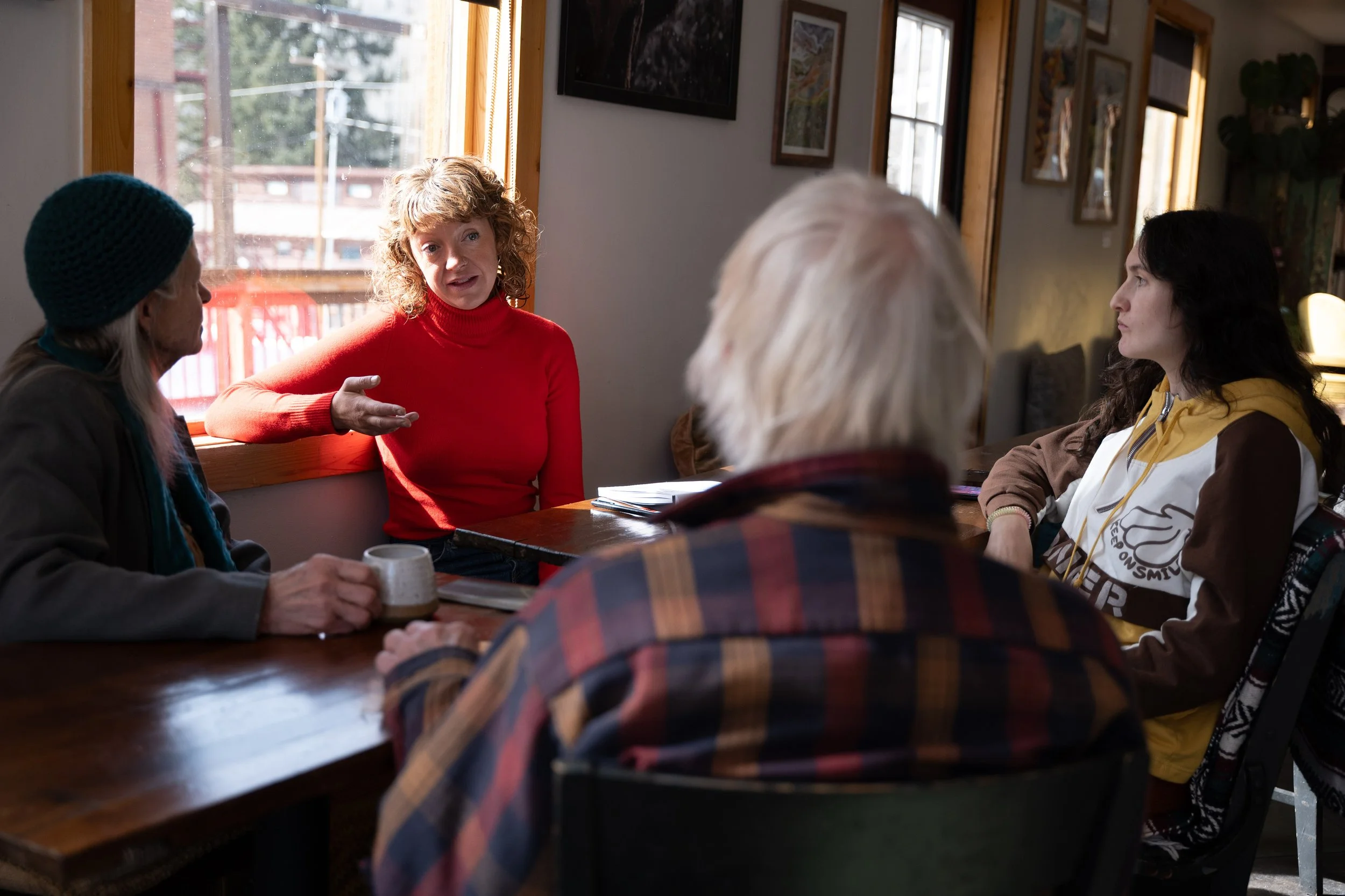 Group of five people sitting around a wooden table having conversation inside a cozy, well-lit room with large windows and artwork on the walls.