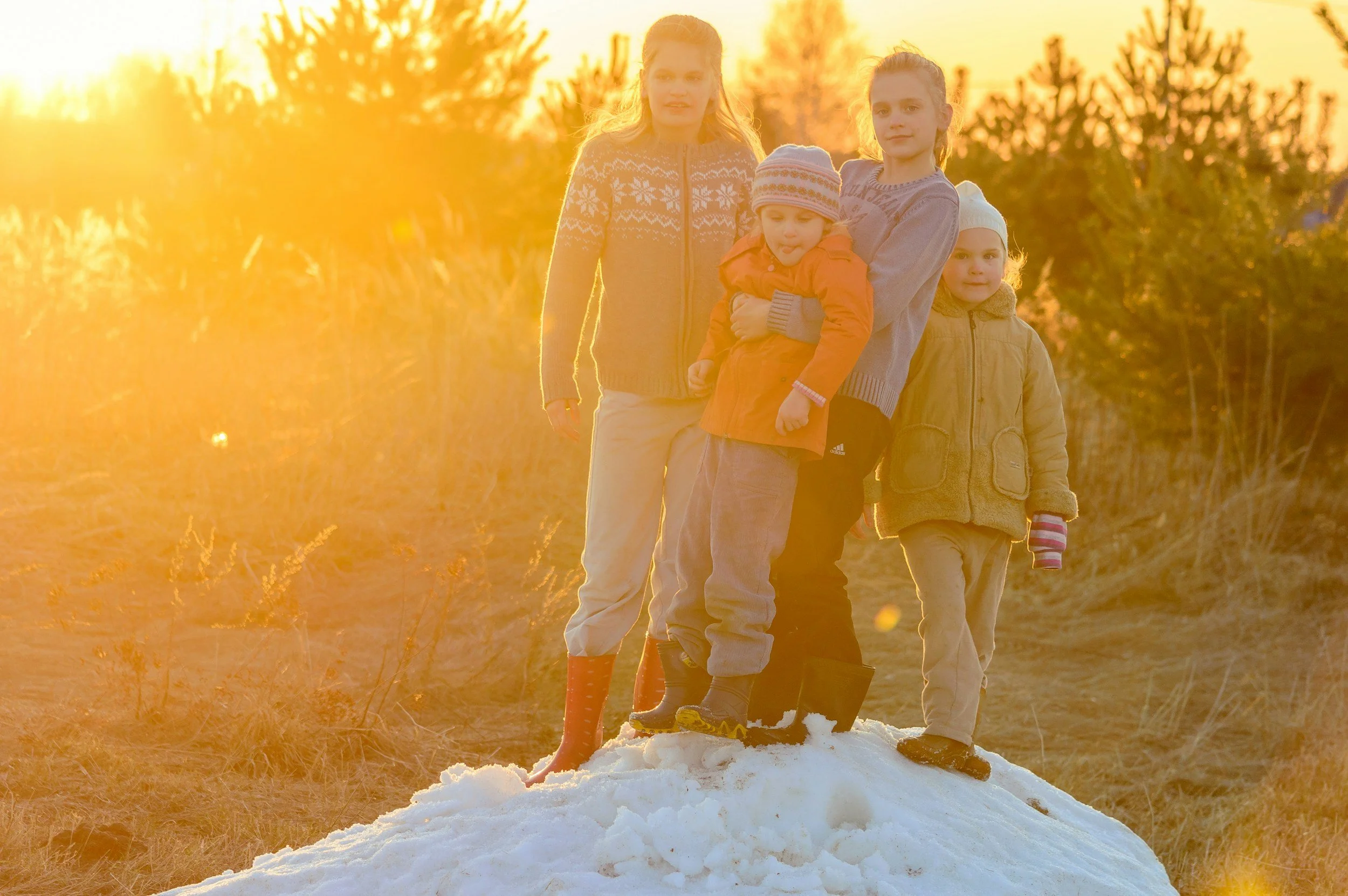 Four children standing on a snow-covered hill during sunset, wearing winter clothing and boots.