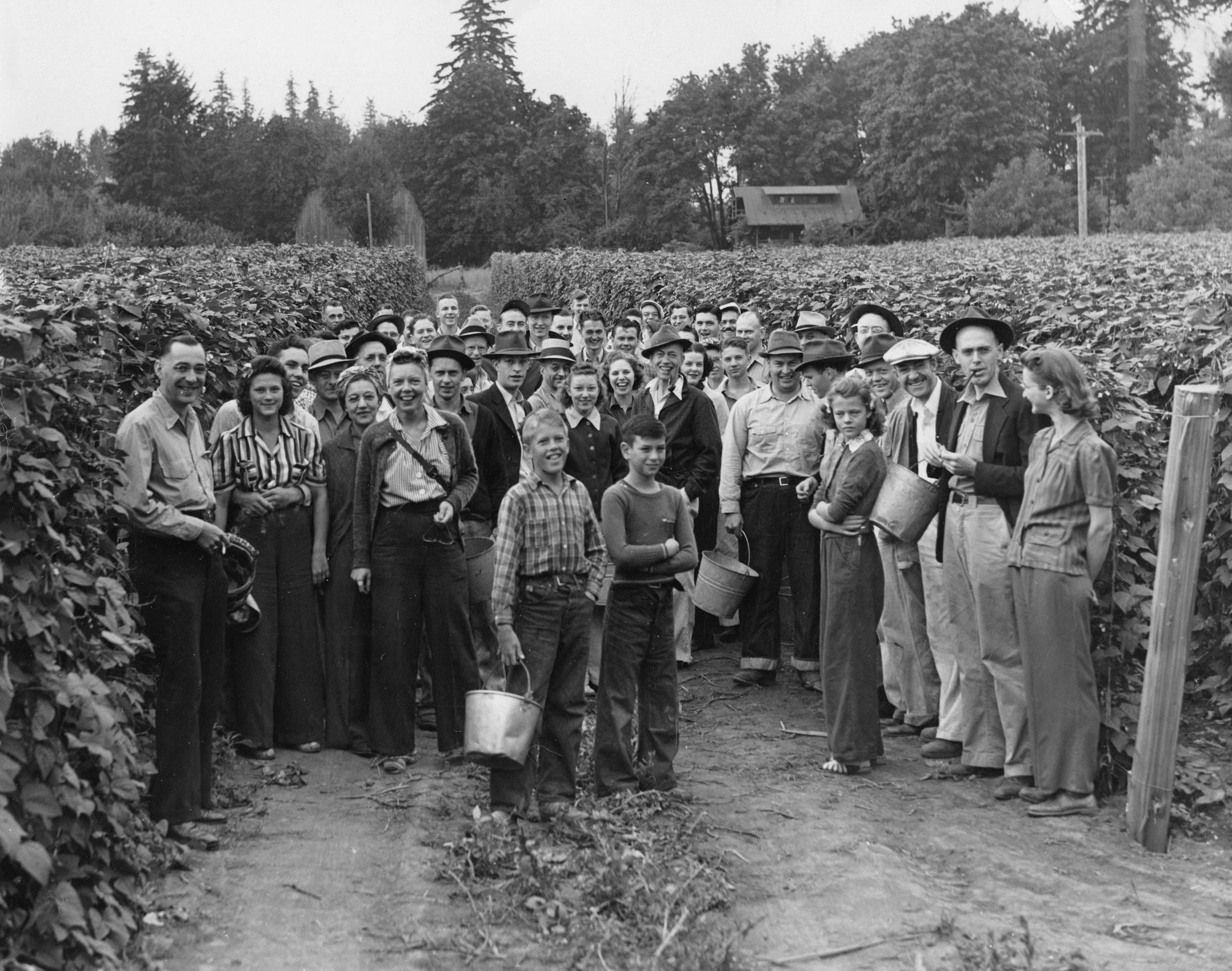 Black and white photo of a group of children and adults standing in a garden or farm, surrounded by rows of plants or crops, with trees and a building in the background.