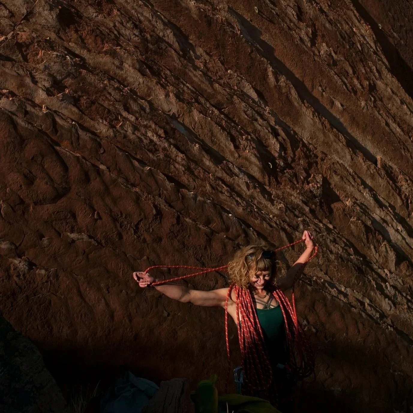 A woman with curly hair is rock climbing inside a cave, using a red rope for safety, with a smile on her face, against a background of a textured brown rocky cave wall.