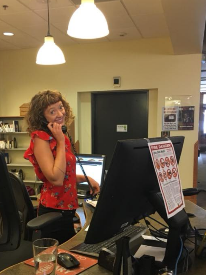 A woman with curly hair wearing a red floral blouse talking on a landline phone at a reception desk in an library environment.