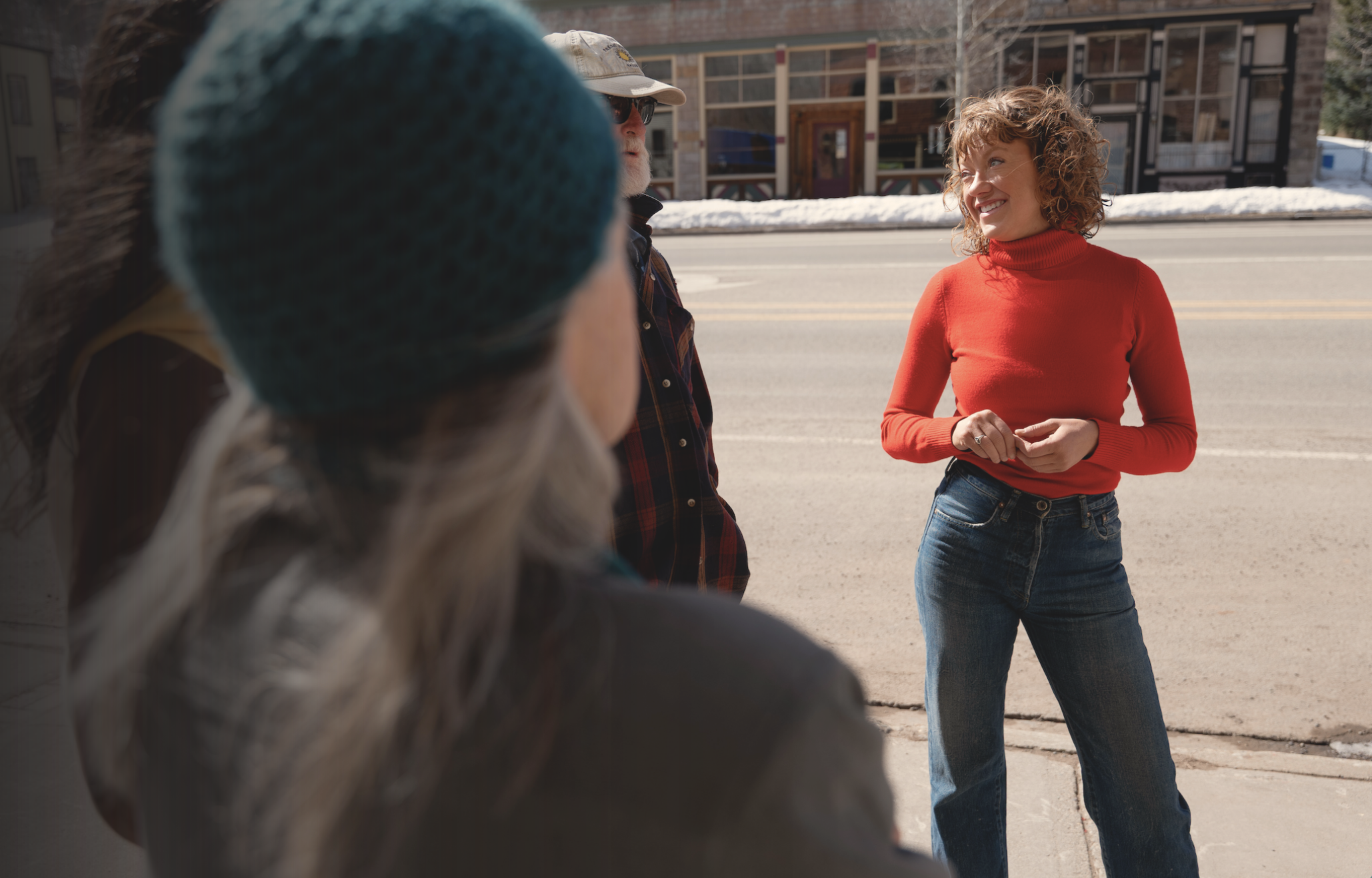 A woman in a red turtleneck sweater smiling and talking with people on a sidewalk in winter, with snow on the ground and a building in the background.