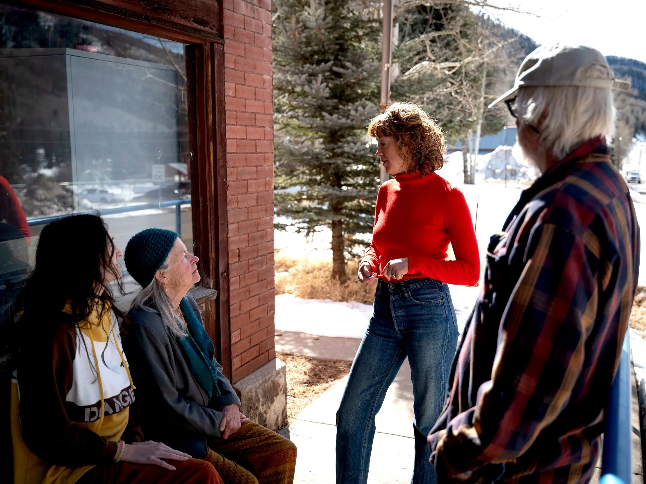 Four women, some elderly, engage in conversation outdoors on a sunny winter day, with one woman standing and the others sitting on a bench beside a brick building, trees, and snow.