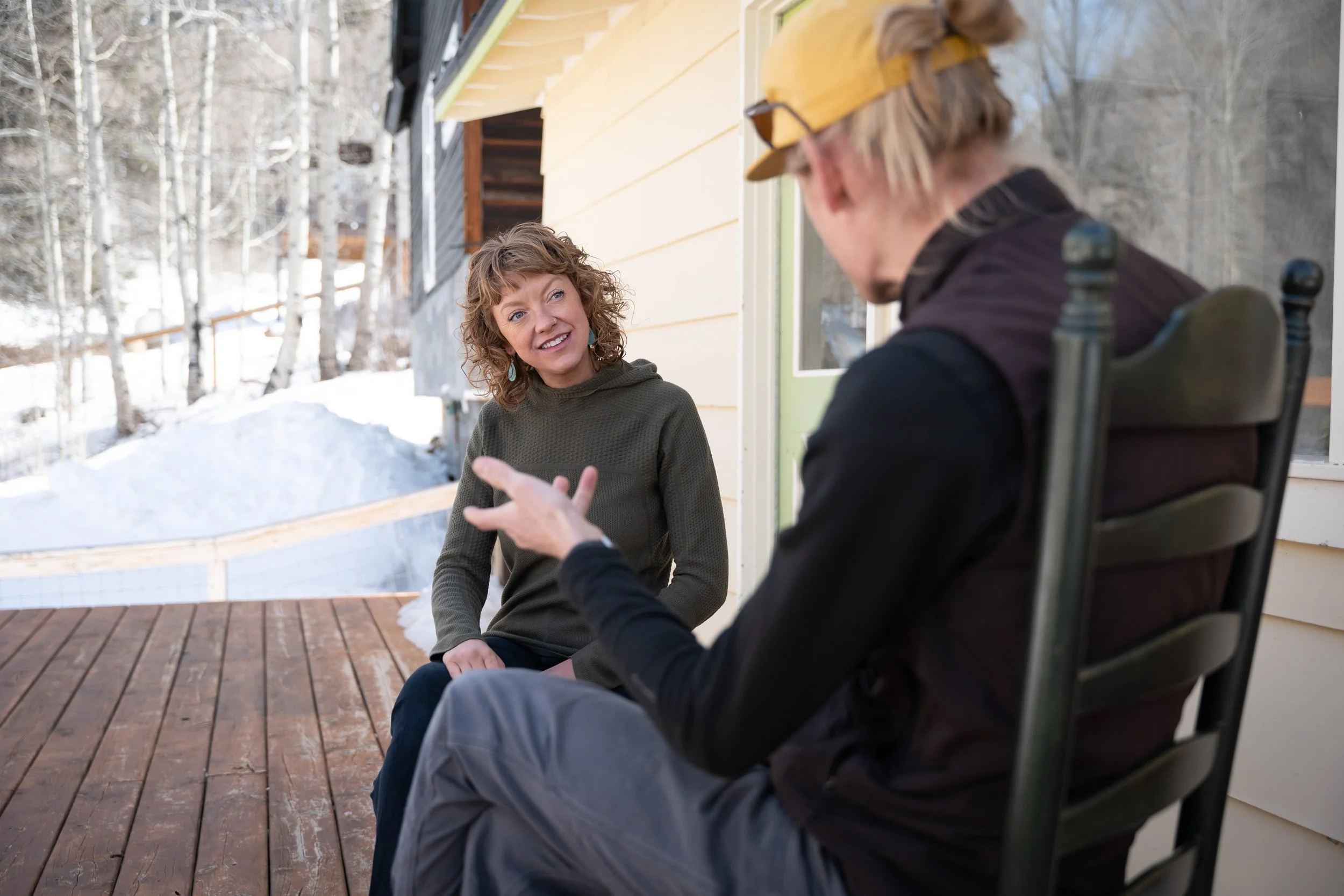 A woman and a man talking outdoors on a snowy porch, with trees in the background. The woman has curly hair and is wearing a dark sweater, smiling at the man. The man has blond hair, a cap, and is sitting in a chair, talking gesture.