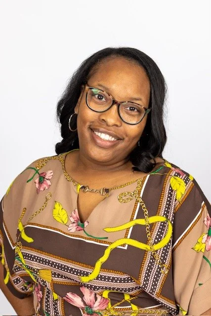 A smiling woman with glasses and earrings wearing a colorful floral and chain pattern blouse, posing against a plain white background.