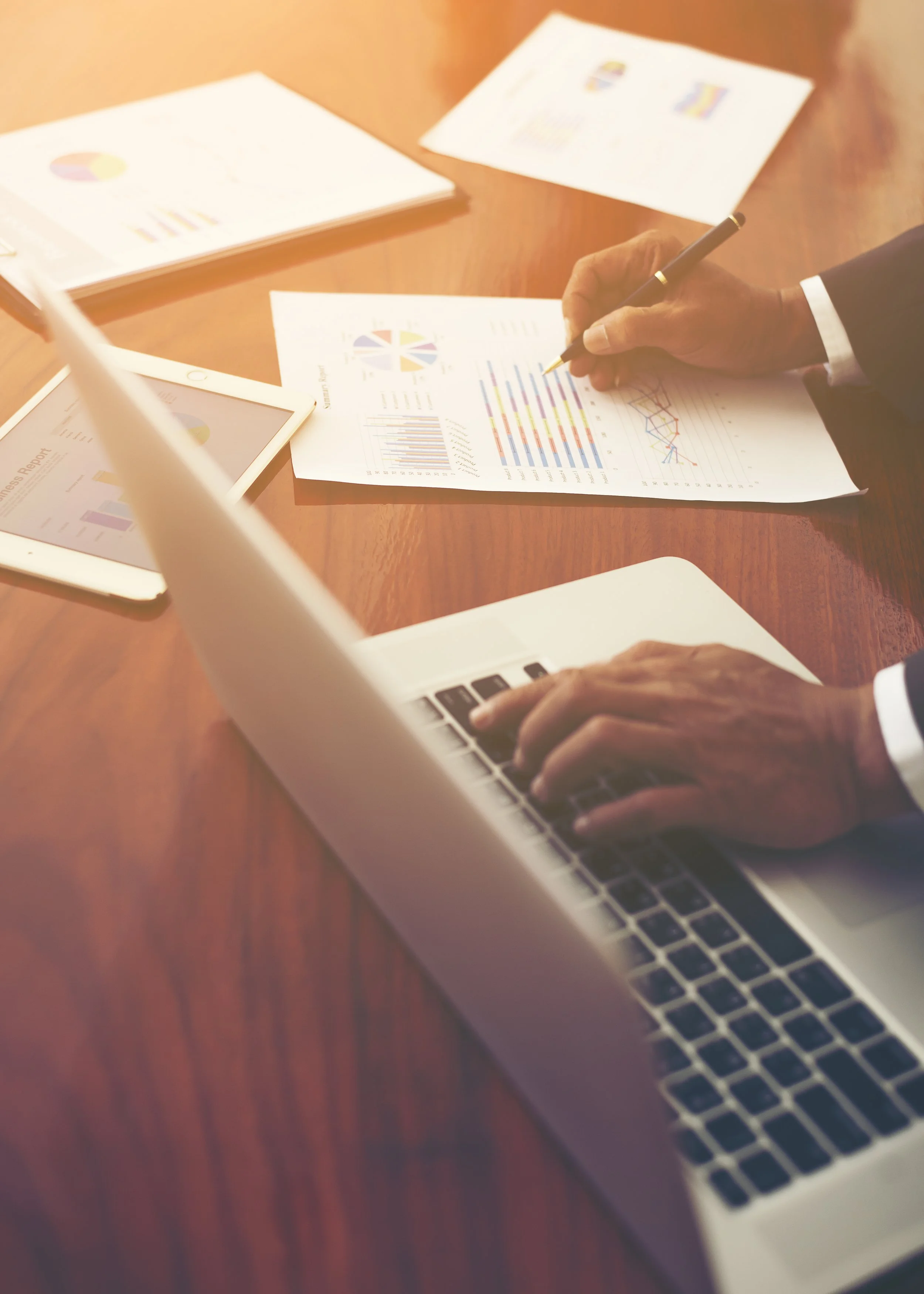 Hands of two people analyzing business reports and graphs on a wooden table, with a laptop, tablet, and printed documents with charts and graphs.