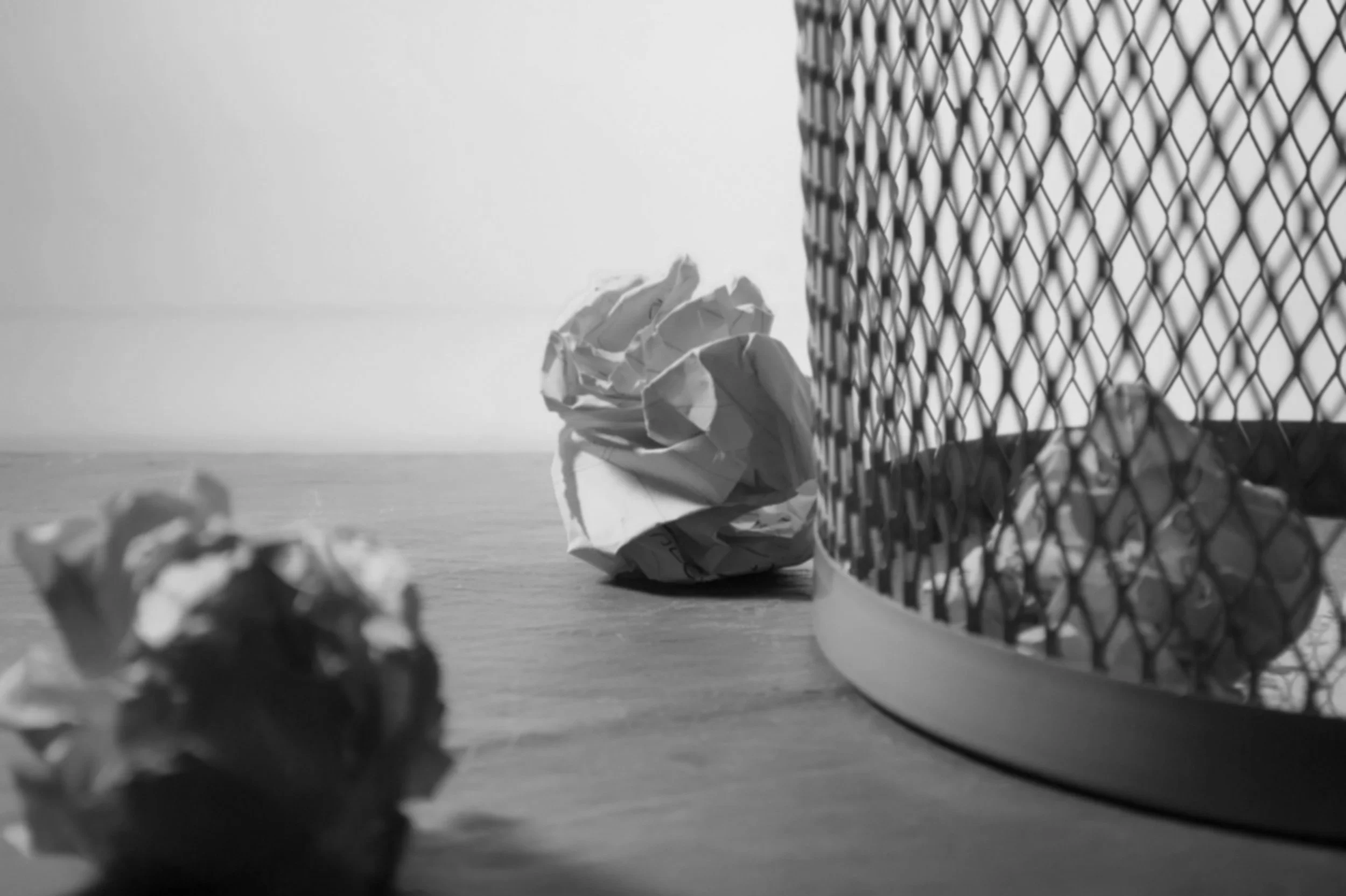 A crumpled piece of paper on a wooden surface, with out-of-focus crumpled papers in the foreground. To the right, there is a metal wire mesh or fence, with the background featuring a body of water and a cloudy sky.