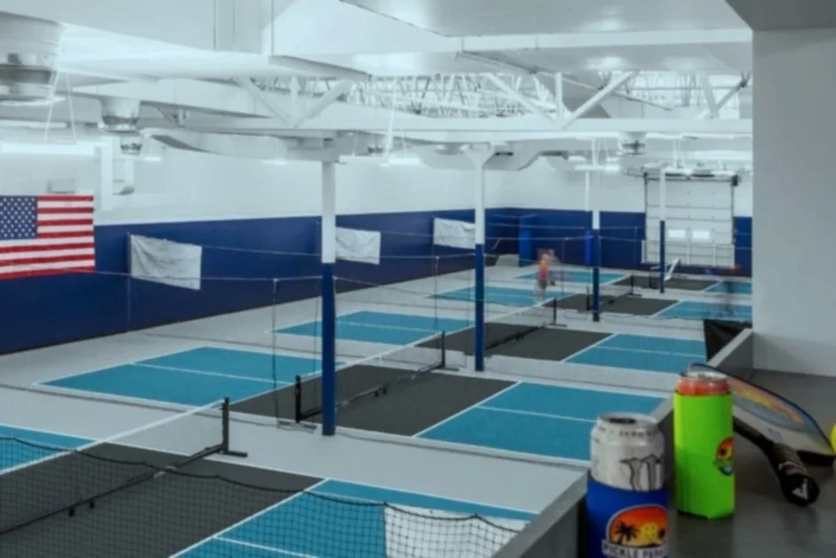 Indoor pickleball courts with blue and gray flooring, barriers, and a person playing in the background. American flag on the wall and drinks on a nearby table.