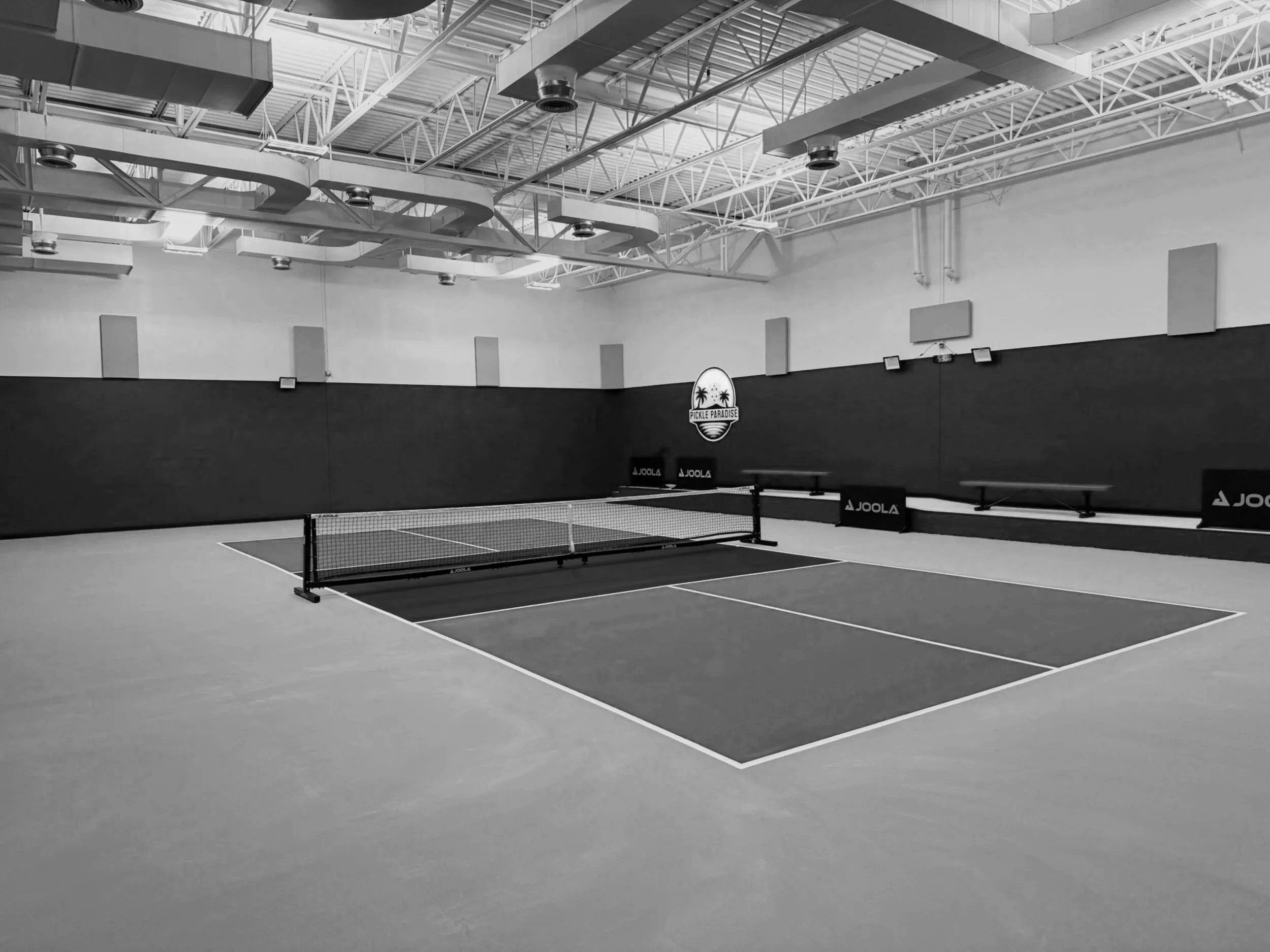 An indoor table tennis court with a net, brand logos on the barriers, and an industrial ceiling with exposed beams and lighting.