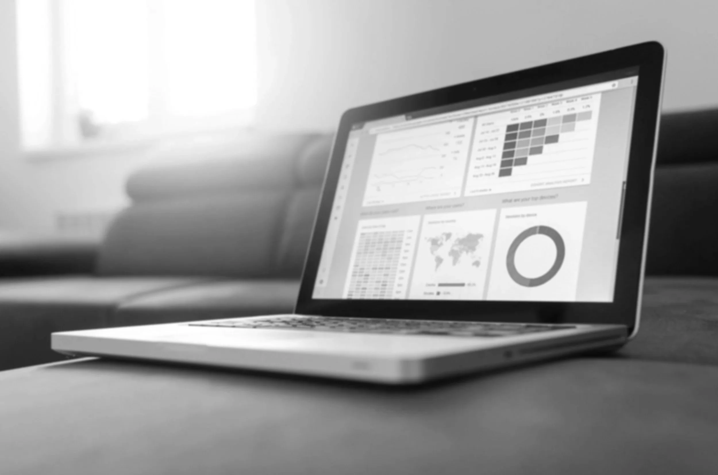 A laptop displaying data charts, graphs, and maps on a desk with a blurred window in the background.