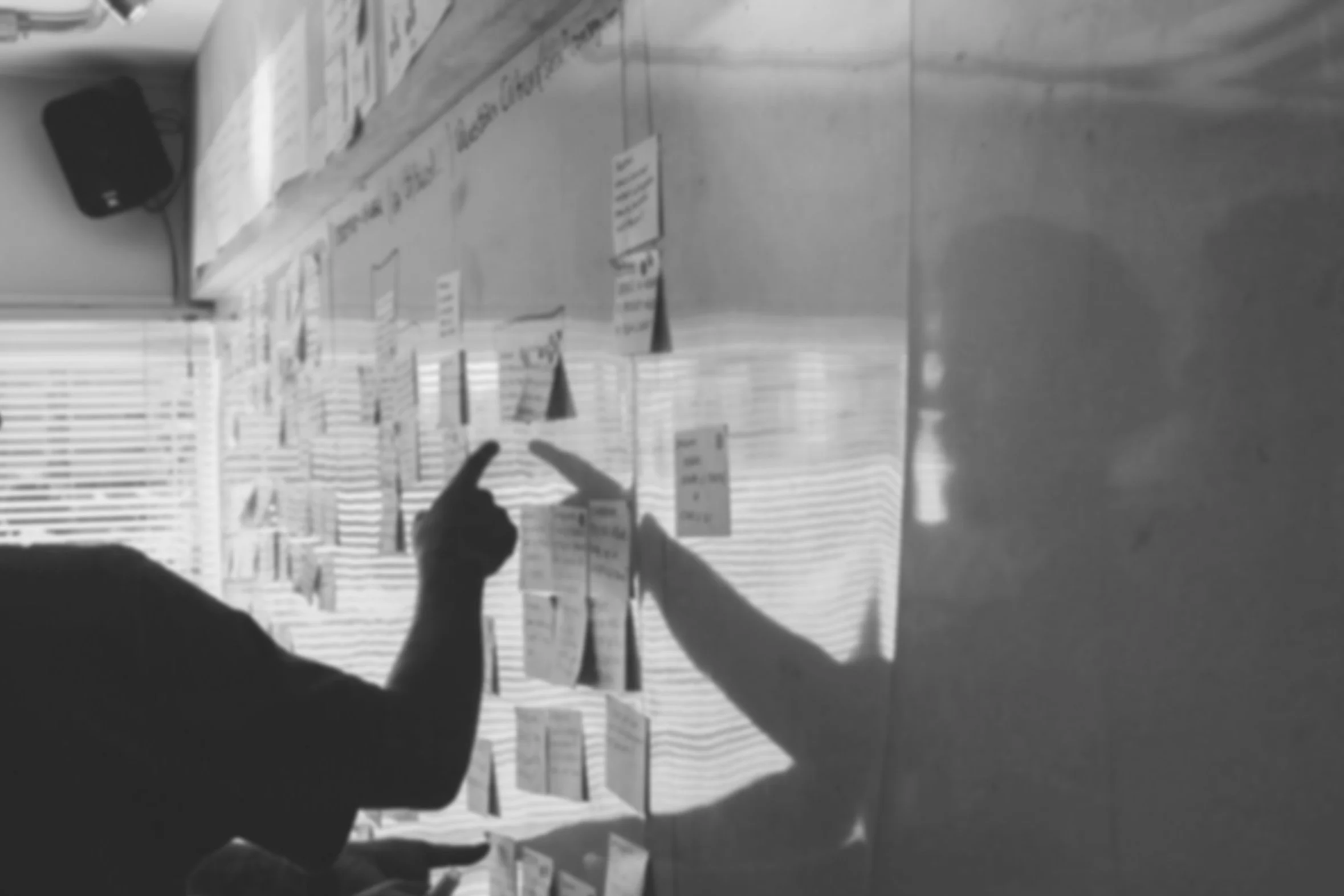 A person taking a photo of a board filled with handwritten notes or labels in a room with fluorescent lighting. The floor appears to be tiled, and a black speaker is mounted on the wall in the background.