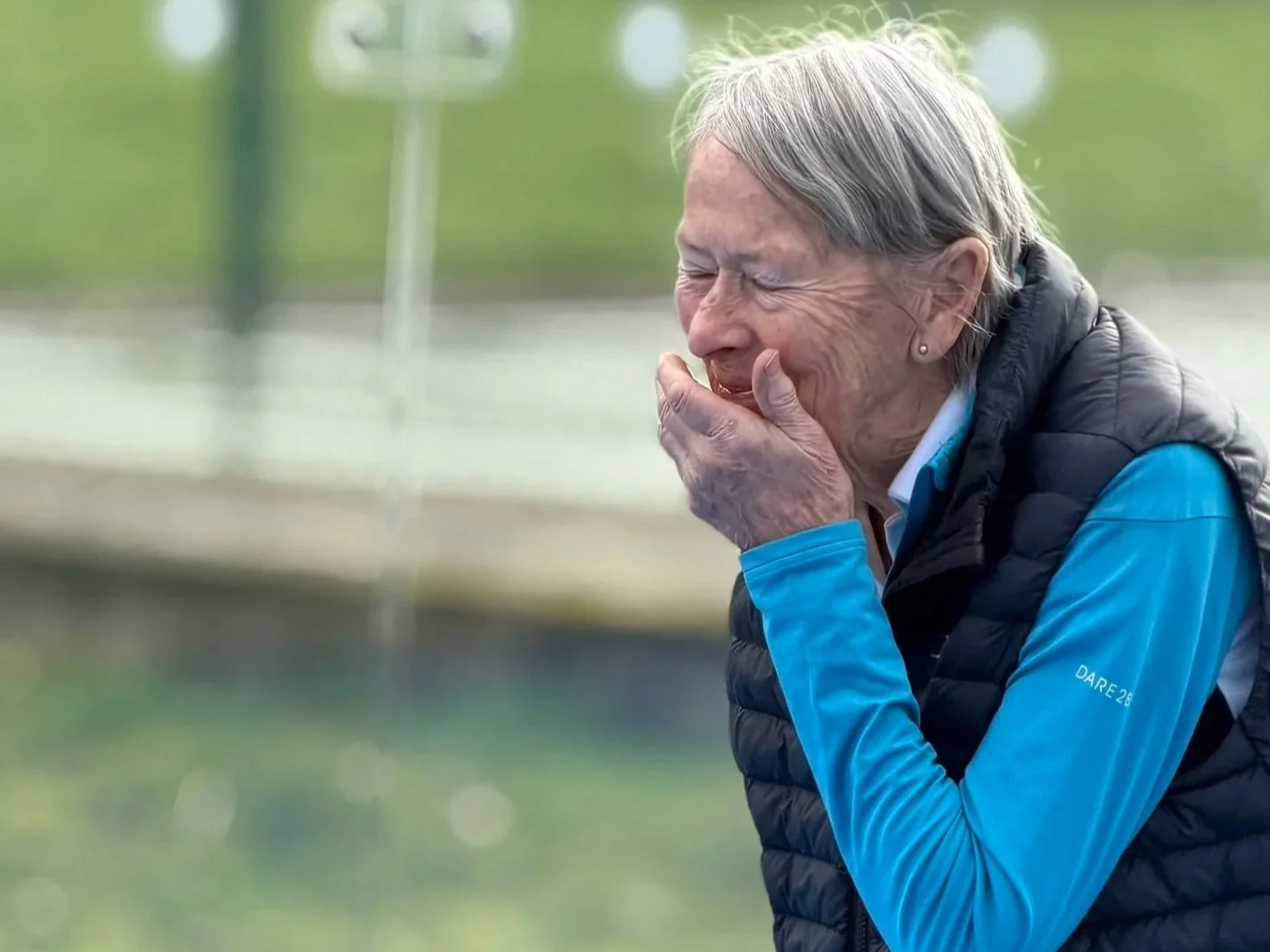 An elderly woman with short gray hair, wearing a blue long-sleeve shirt and black puffer vest, is laughing while covering her mouth with her hand outdoors.