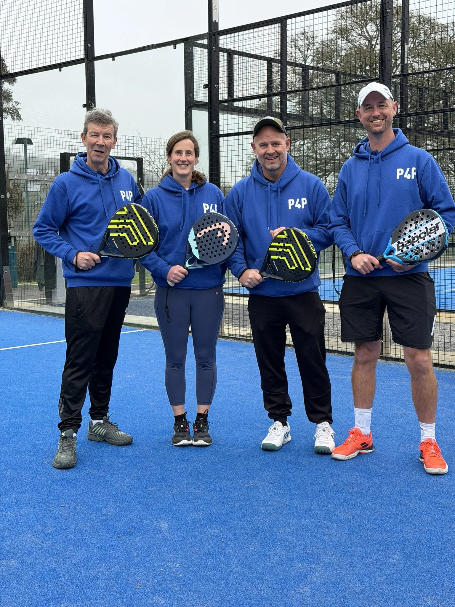 Four people standing on a blue padel court, holding padel rackets, dressed in blue hoodies with 'P4P' logo, smiling at the camera, enclosed by a black wire fence with trees in the background.