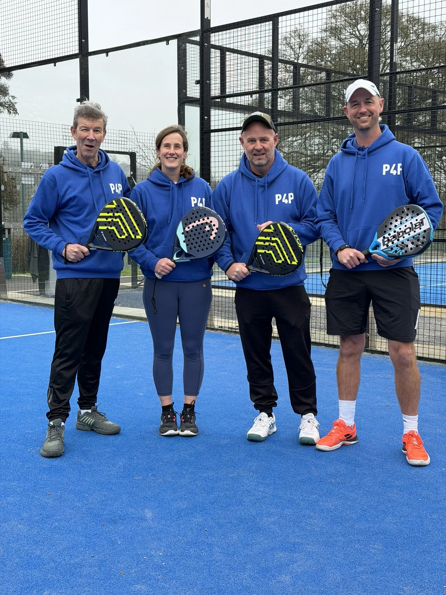 Four people standing on a blue padel court holding padel rackets, with a fenced background and trees visible. They are wearing blue hoodies with P4P printed on them, smiling, and dressed in athletic shoes.