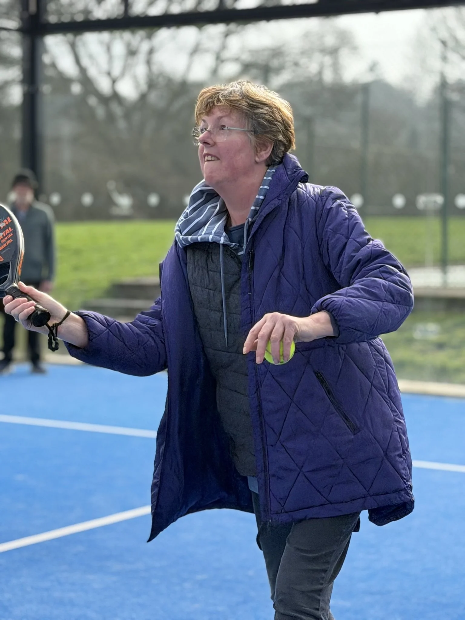 Woman in a purple jacket playing tennis on a blue court outdoors, holding a tennis ball and racket.