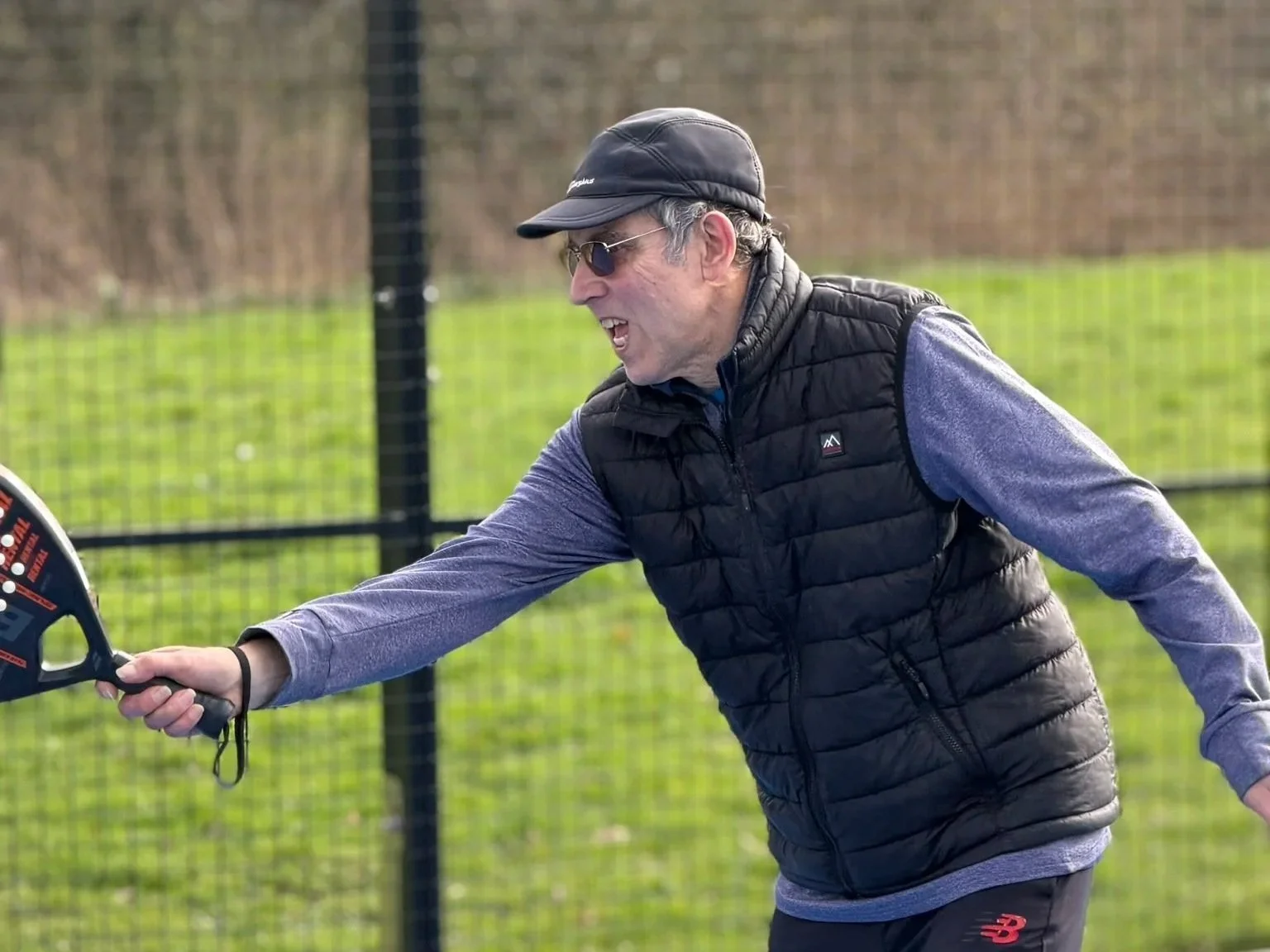 Man wearing sunglasses, black cap, black vest over purple long-sleeve shirt, playing pickleball outdoors, reaching to hit the ball with a paddle.