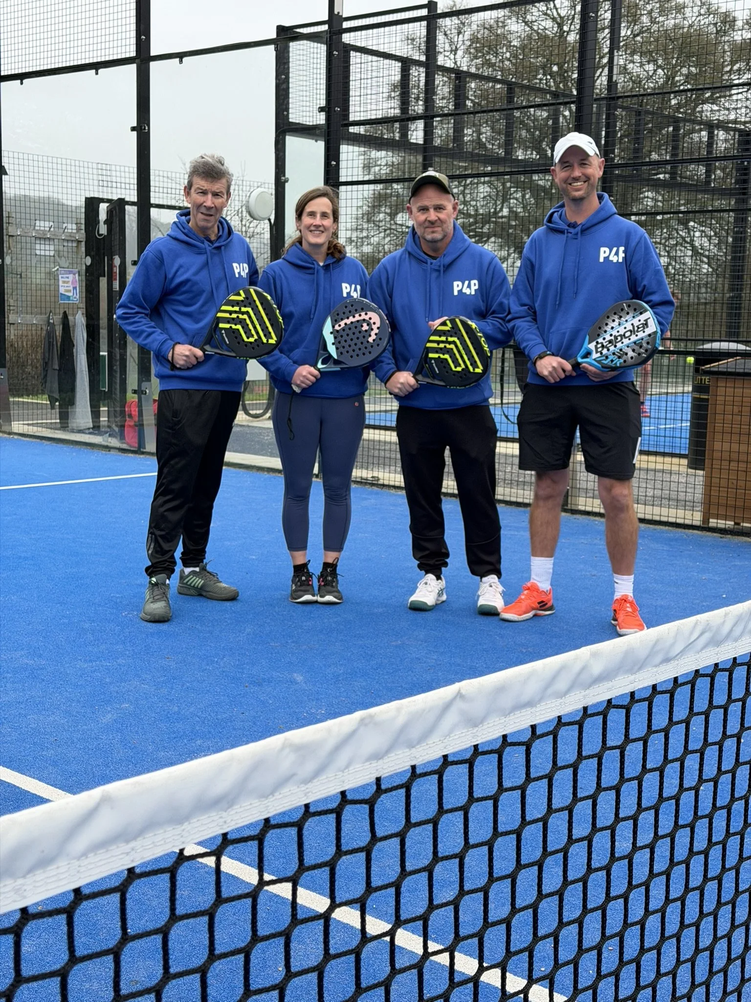 Four people standing on a blue padel tennis court holding paddles, with a black wire fence in the foreground and trees in the background. They are wearing blue hoodies with P4L printed on them.