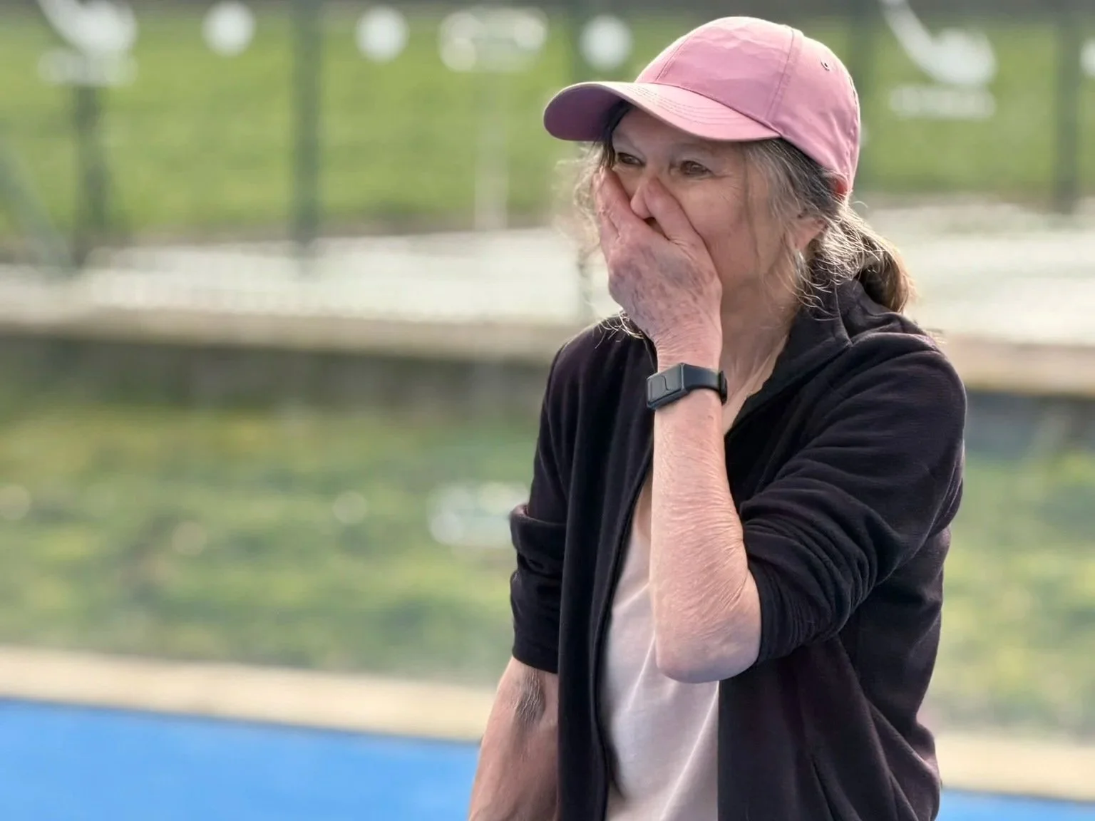 An elderly woman in a pink cap and black jacket covering her mouth, appearing emotional or surprised, outdoors near a tennis court.