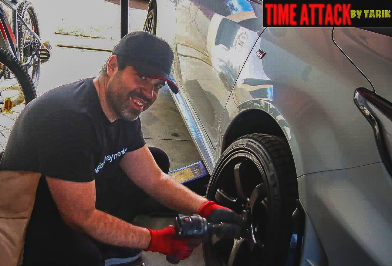 A man wearing a black cap, black t-shirt, and red gloves is changing a tire on a silver car in a garage. The garage contains a bicycle and tools in the background. There is a logo "TIME ATTACK" by Tarik in the top right corner.
