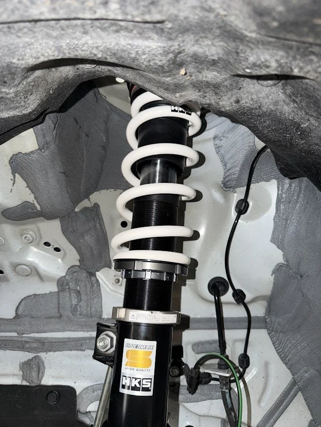 Close-up view of a car's suspension coilover, with a black shock absorber and white coil spring, installed in the wheel well.