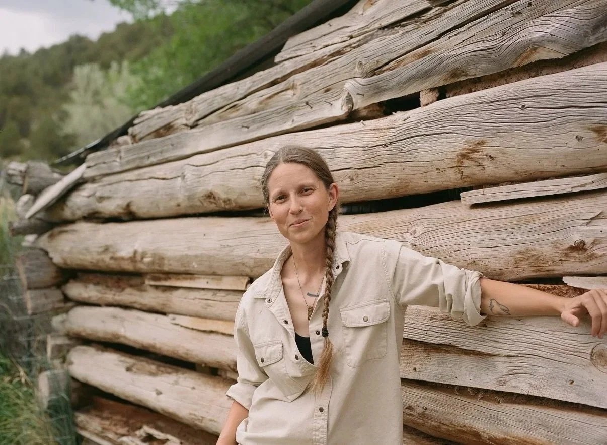 A woman with a braid and tattoo on her arm, wearing a beige shirt, leaning against a rustic wooden log cabin outdoors in a natural setting.