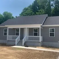Front view of a small, single-story house with gray siding, white trim, and a covered porch with white railings and stairs.
