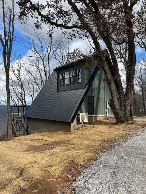 A raised green house with a metal roof, situated on a sloped yard with a gravel path, near leafless trees under a partly cloudy sky.
